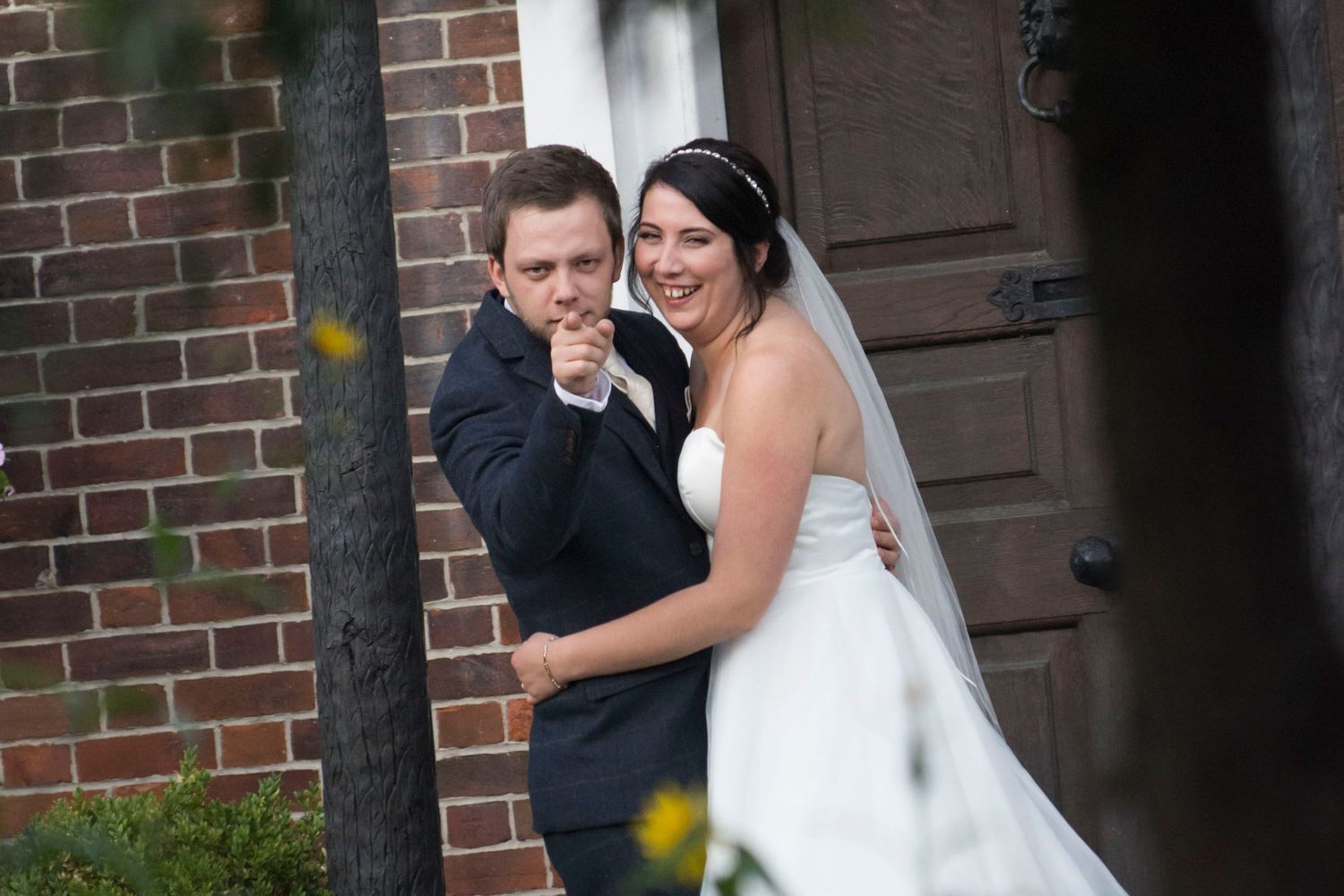 Smiling groom playfully pointing at camera while bride in white gown laughs beside him near brick building.