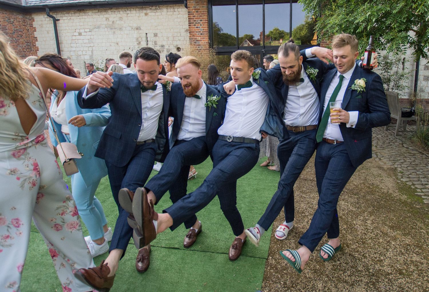 Groosmen in navy suits doing a fun high-kick dance pose outdoors at wedding reception.