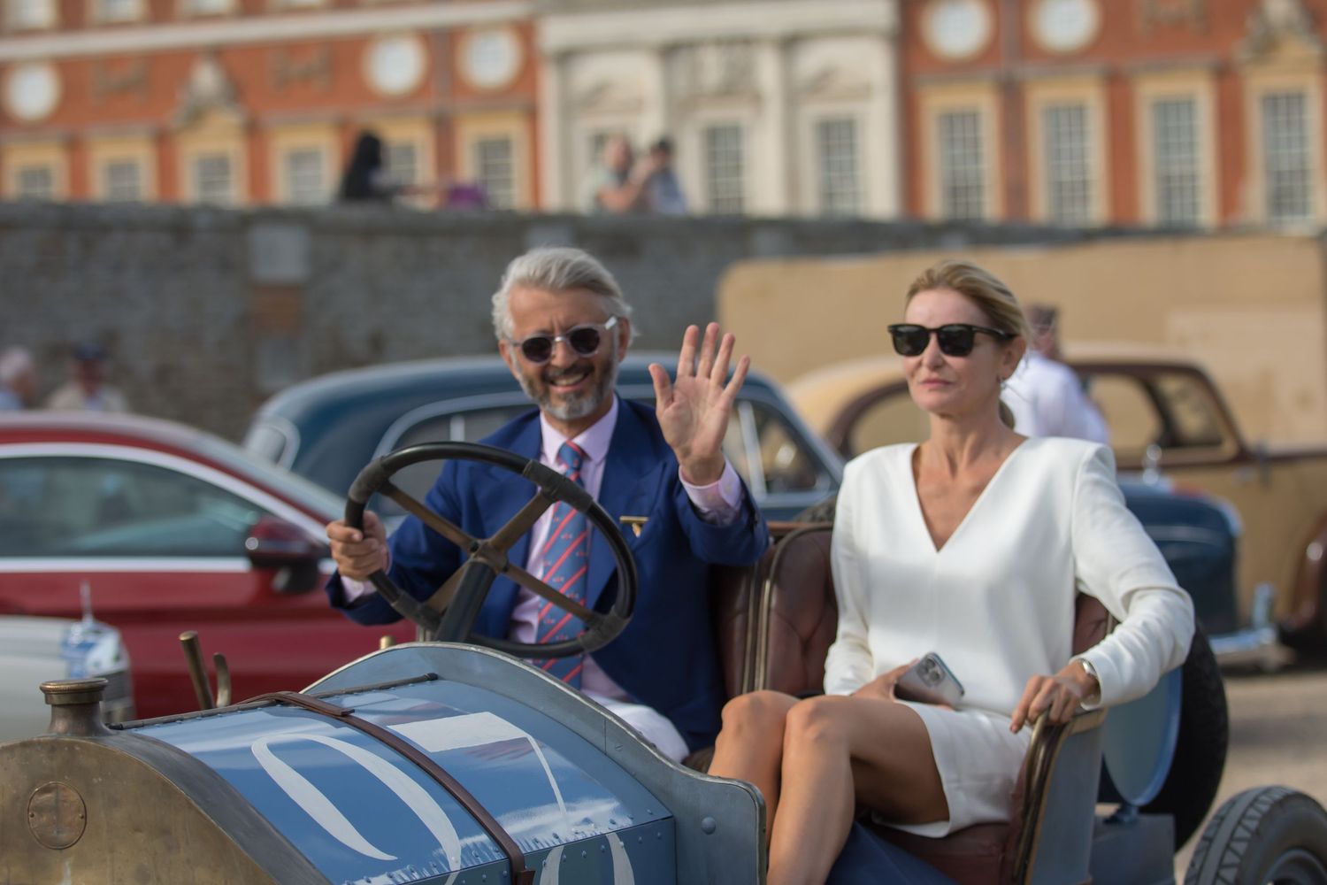 Stepane Durraq  waves from a vintage racing car  leaving Concours of elagance at Hampton Court Palace whilst Katarina Kyvalova holds on