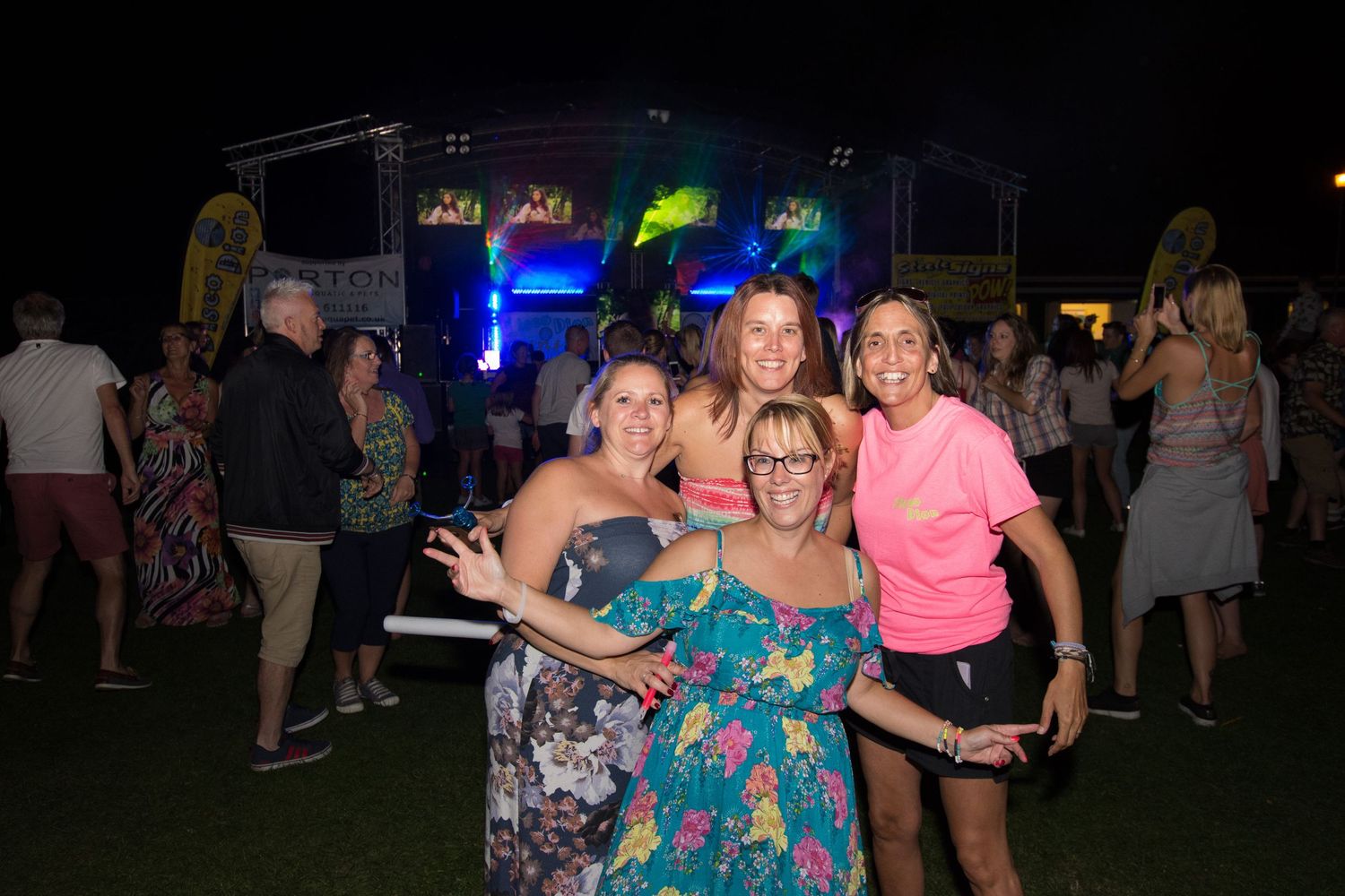 Four women pose together smiling at an outdoor night concert event with colorful stage lights in the background.