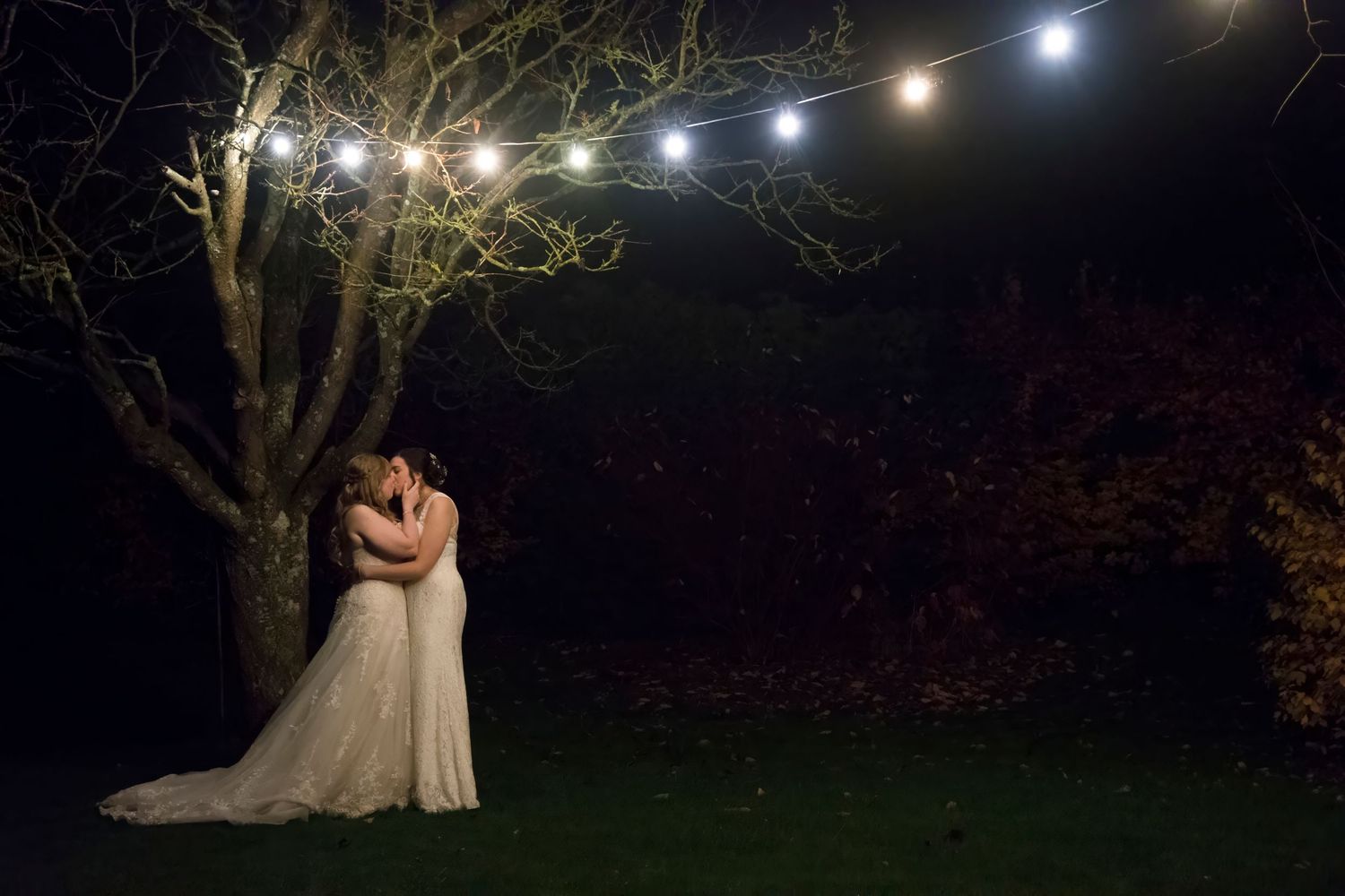 Two brides in white gowns sharing a romantic kiss under glowing string lights in a dark garden at night.