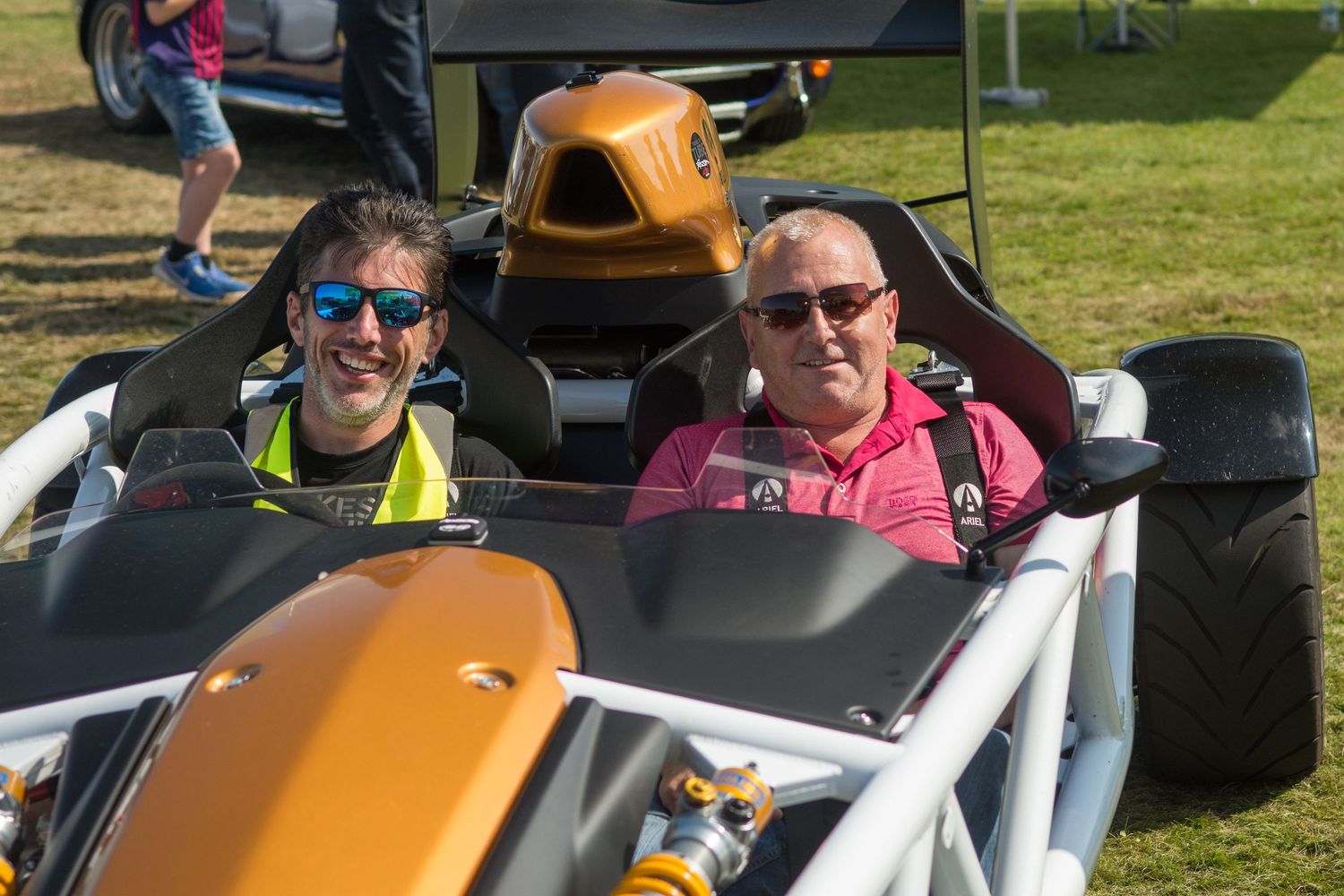 Two men smiling in an Ariel Atom sports car at an the british motor show on a sunny day.