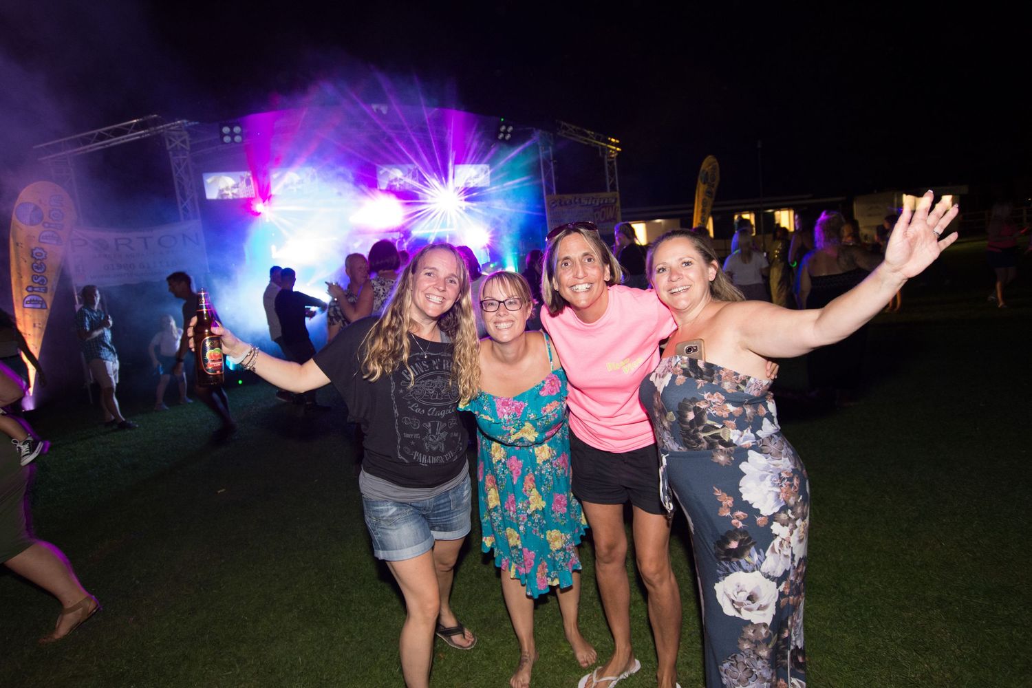 Four women posing happily at an outdoor concert at night with colorful stage lights in the background.