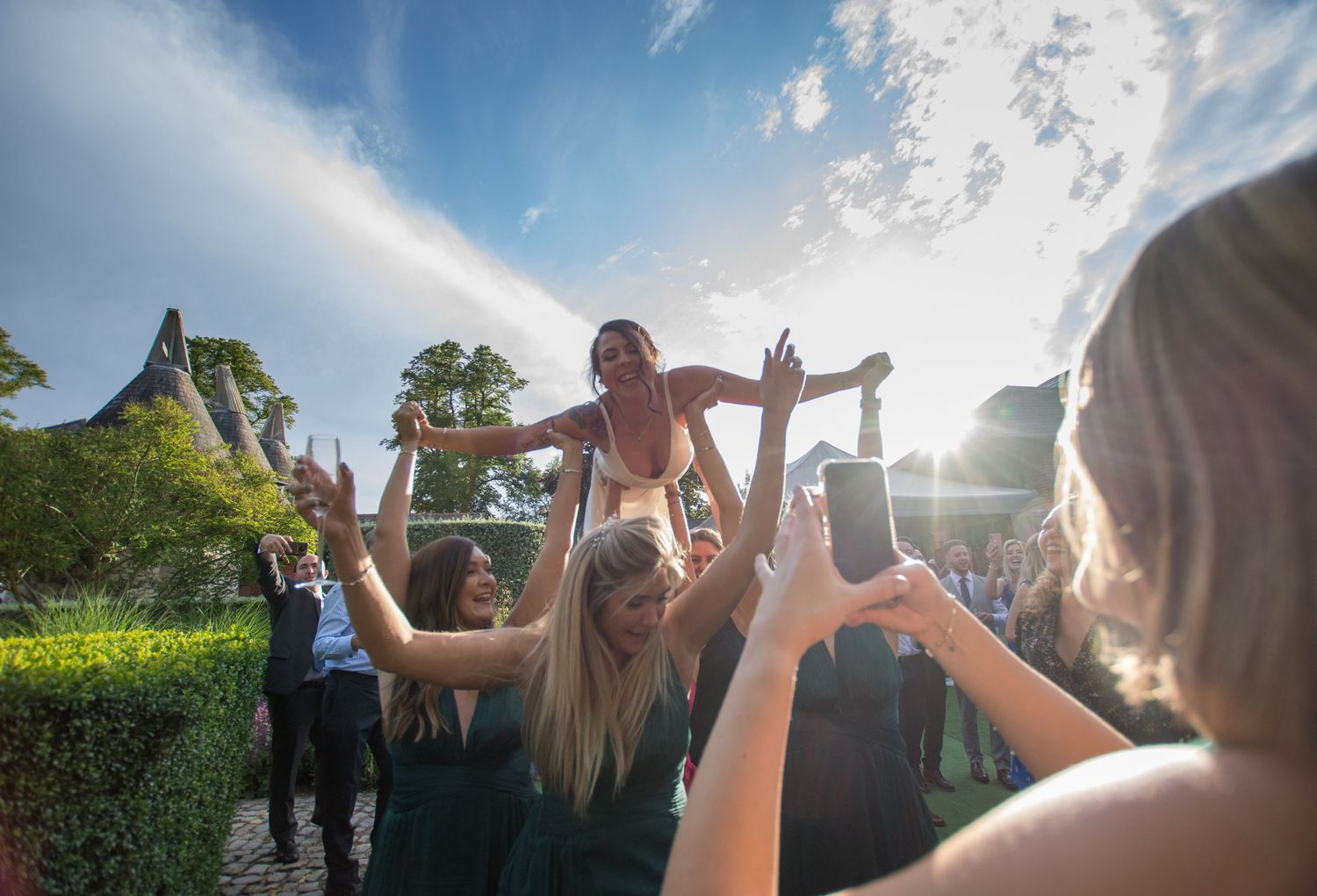 Joyful Bride lifted by her bridesmaids above crowd with arms raised in sunny outdoor celebration.