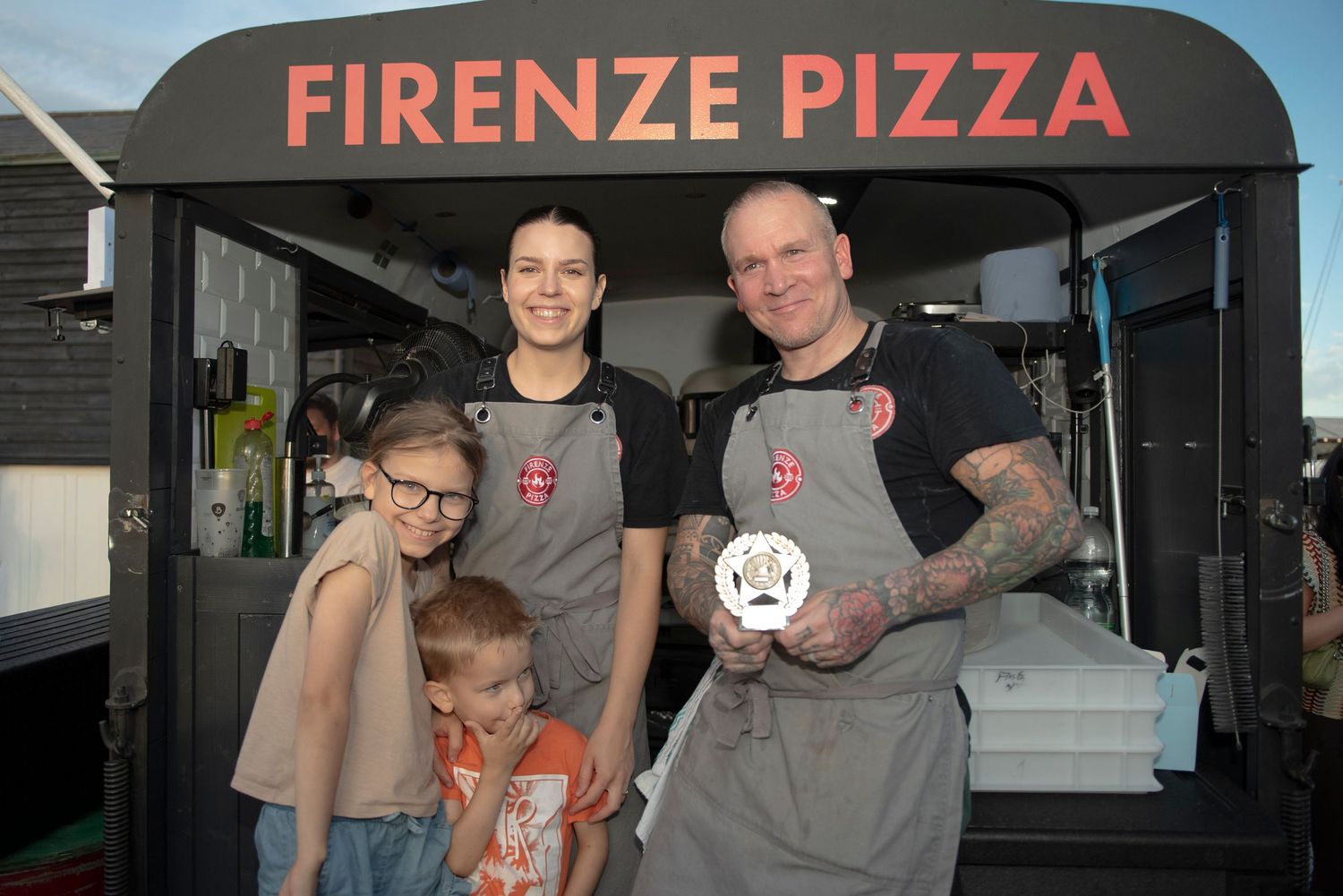 Two adults and two children pose smiling in gray aprons in front of a Firenze Pizza food truck after winning a prize at Essex Pizza Festival