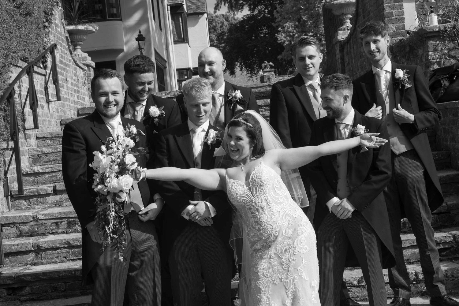 Black and white photo of smiling bride in lace gown photobombing the posing  groom and groomsmen on stone steps.
