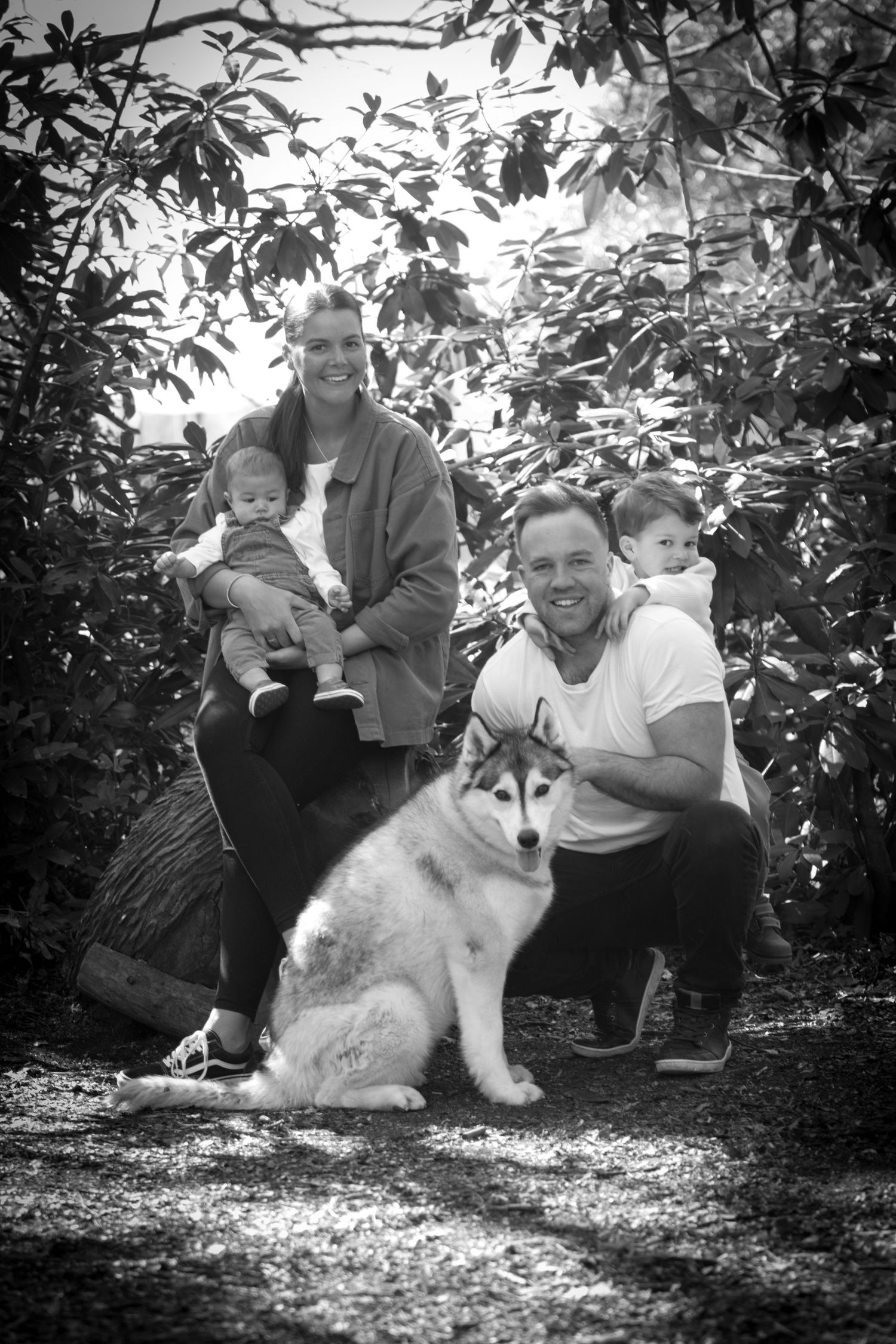 Black and white family portrait with two adults, two babies, and a Husky dog outdoors among lush foliage.