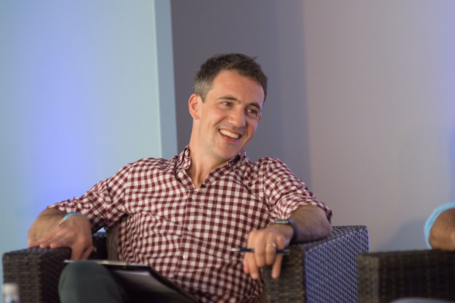 Smiling Joe Crowely in red checkered shirt relaxing in wicker chair during a casual panel discussion.