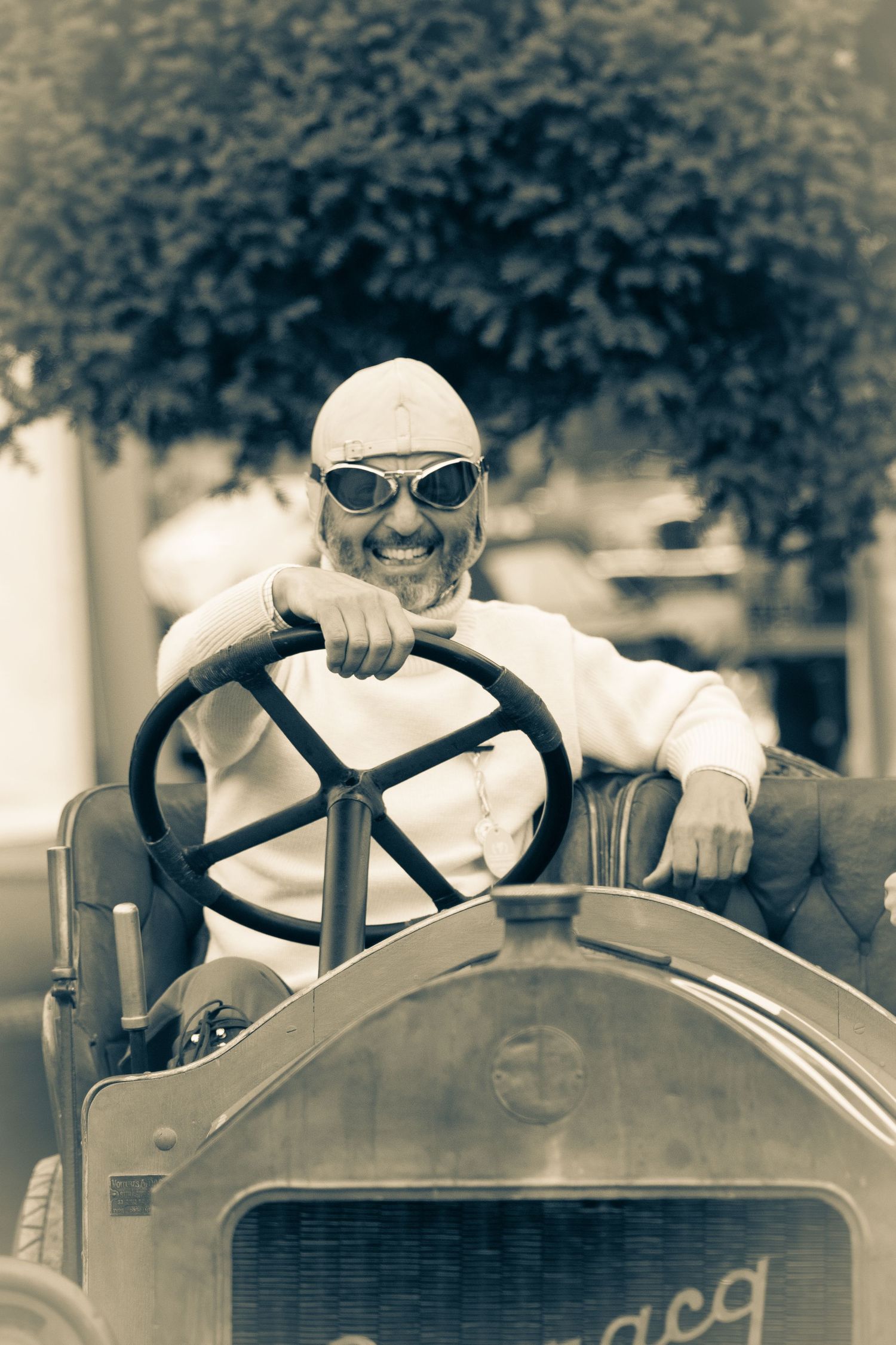 A Stephane Durraq in vintage racing gear drives a classic antique car in a sepia-toned photo.