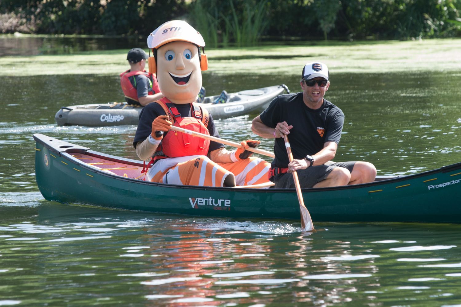 Man paddling a canoe with a cheerful mascot character wearing a life jacket on a calm river.