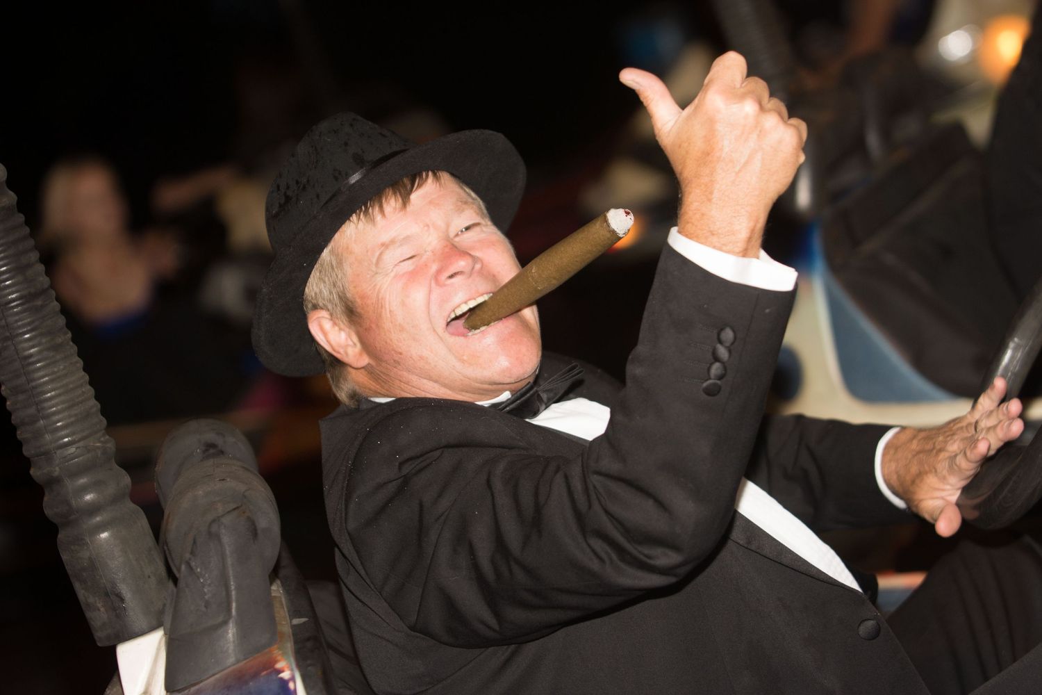 A man in a black tuxedo and hat smokes a cigar and gives a thumbs up at a formal event.
