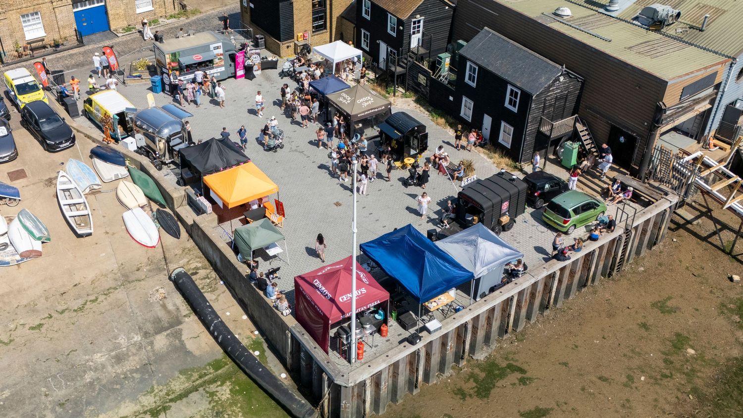Aerial view of a busy Essex Pizza Festival with colorful tents and crowds near waterfront buildings on a sunny day.