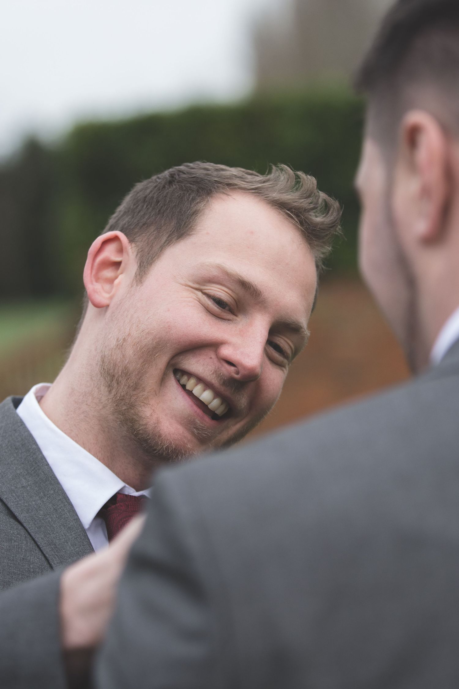 Smiling groomsman in grey suit sharing a joyful moment with another guest outdoors.