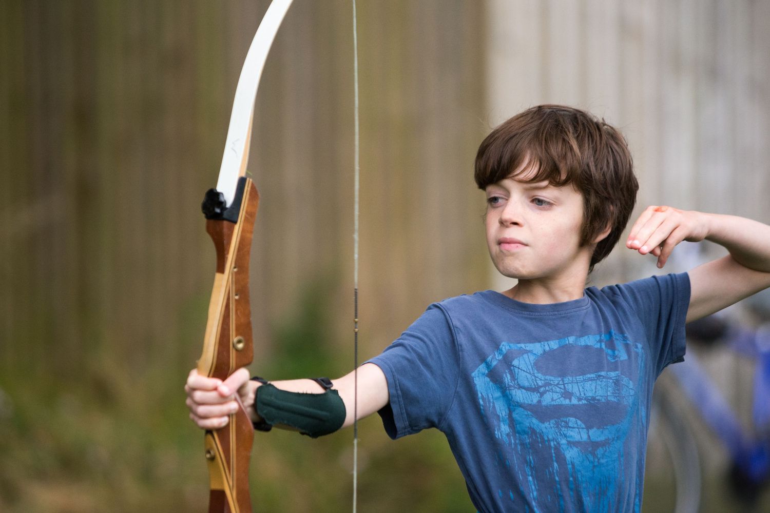 Young boy practicing archery, drawing a wooden bow with focus and determination outdoors.