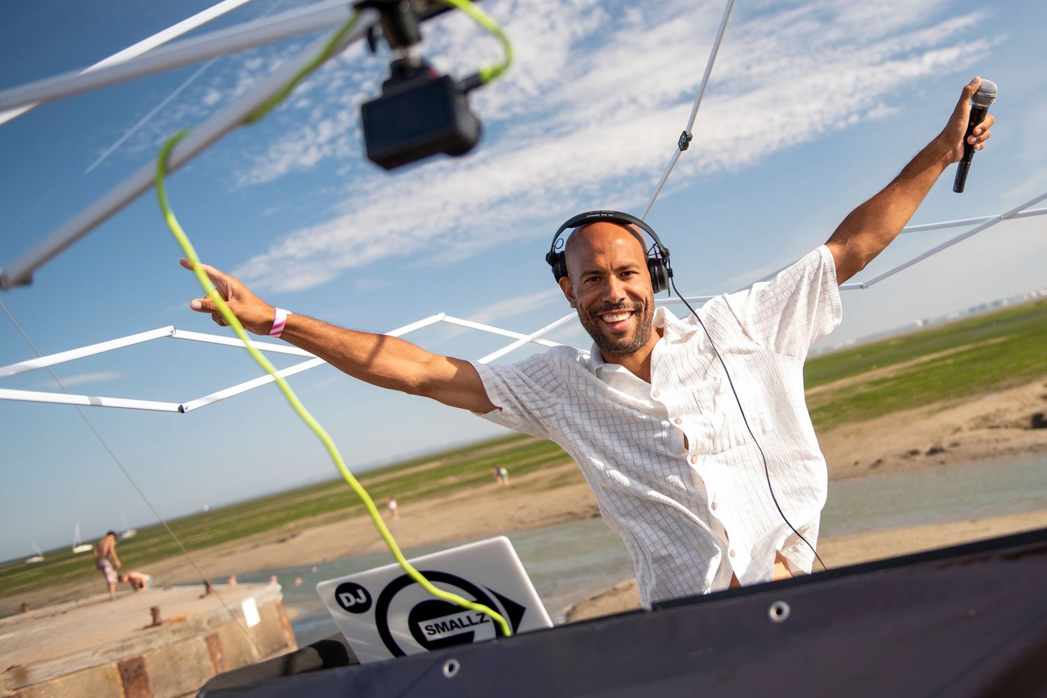 A smiling DJ with headphones raises his arms behind decks at Essex Pizza festival under a blue sky.