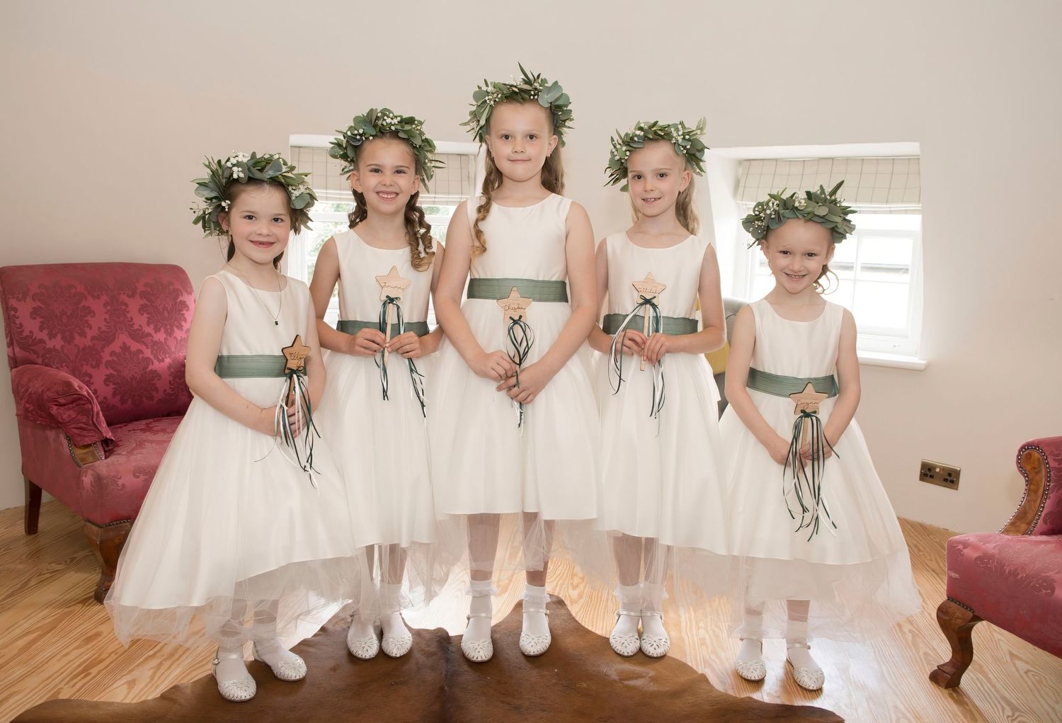 Five flower girls in white dresses with green sashes and floral crowns pose together.