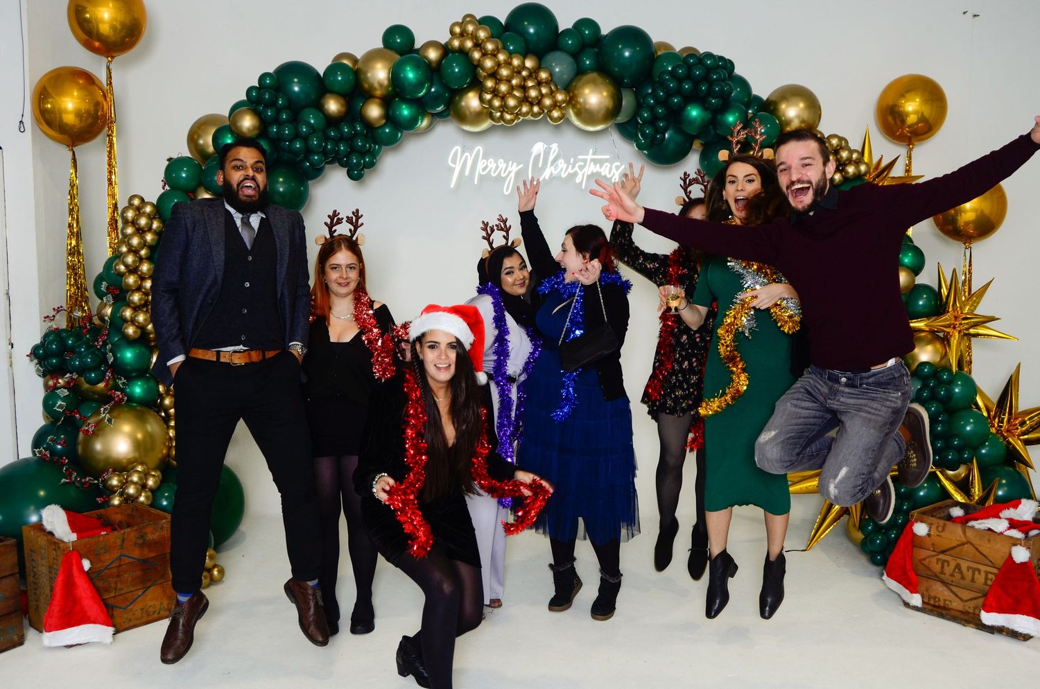 Group of seven people jumping and celebrating in front of a festive Christmas balloon arch.