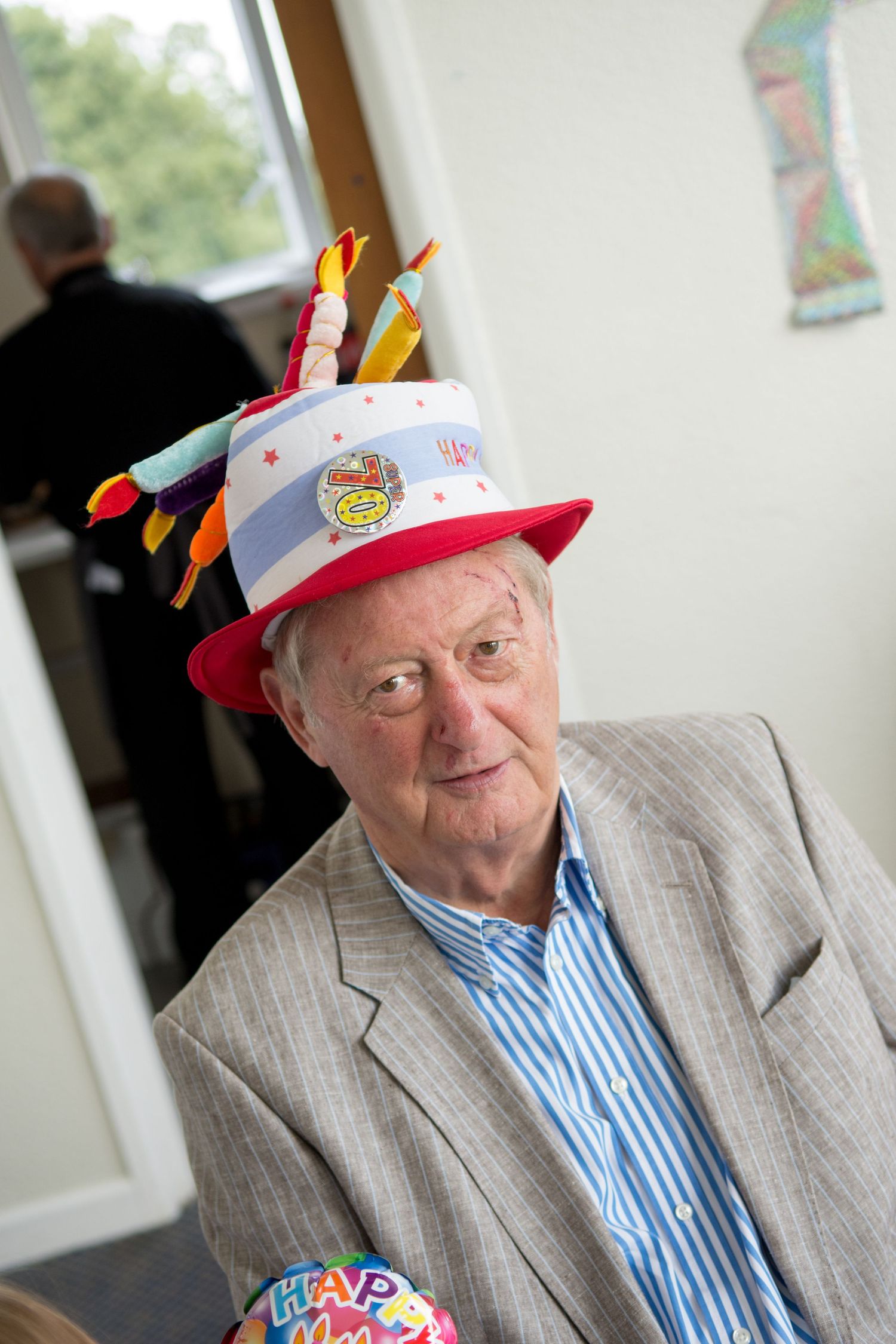 Elderly man wearing a colorful birthday cake novelty hat with candles at a party celebration indoors.