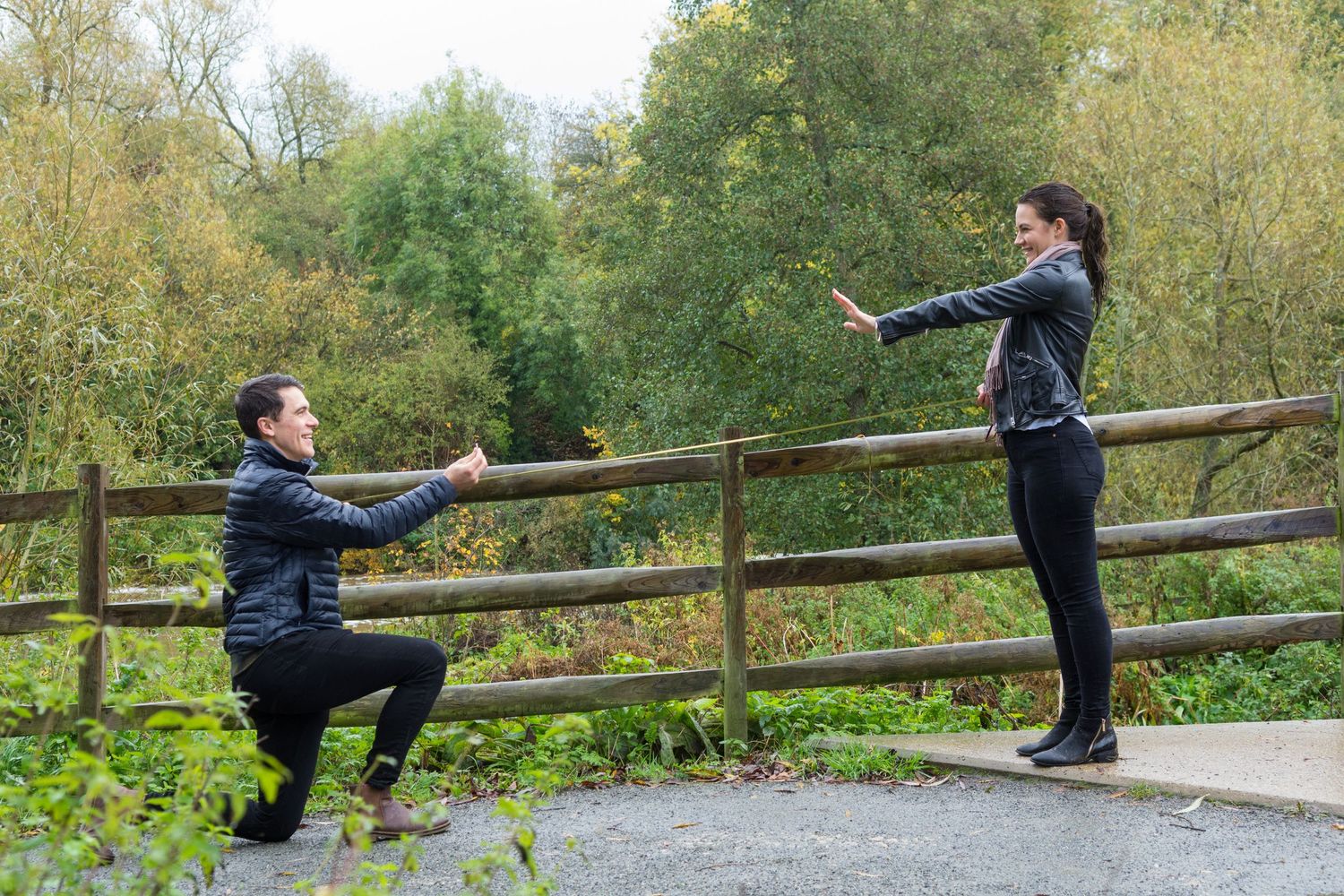 Man kneeling on one knee proposing to a woman outdoors near a wooden fence with lush green trees in background.