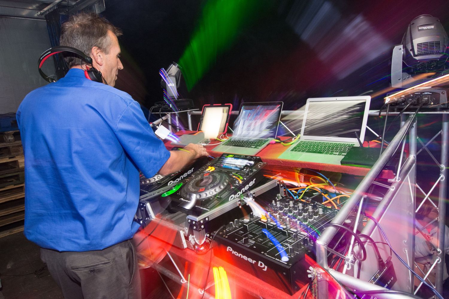 DJ mixing music at a professional setup with laptops and controllers under colorful stage lights.