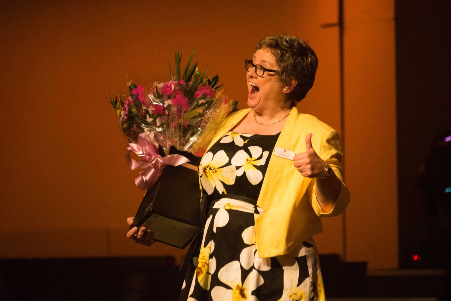 Excited woman in yellow jacket holding flowers and giving thumbs up on stage at awards event.