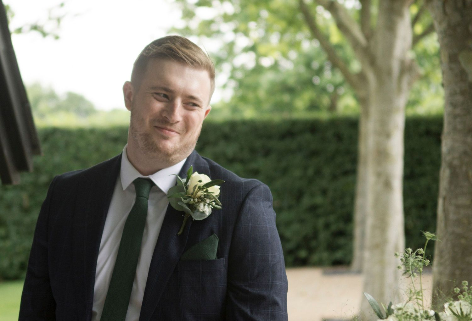 Handsome groom smiles wearing navy suit with green tie and boutonniere outdoors.
