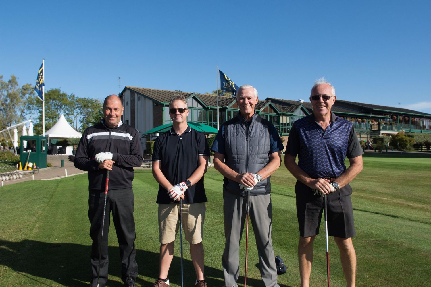 Four men posing with golf clubs in front of a golf clubhouse on a sunny day.