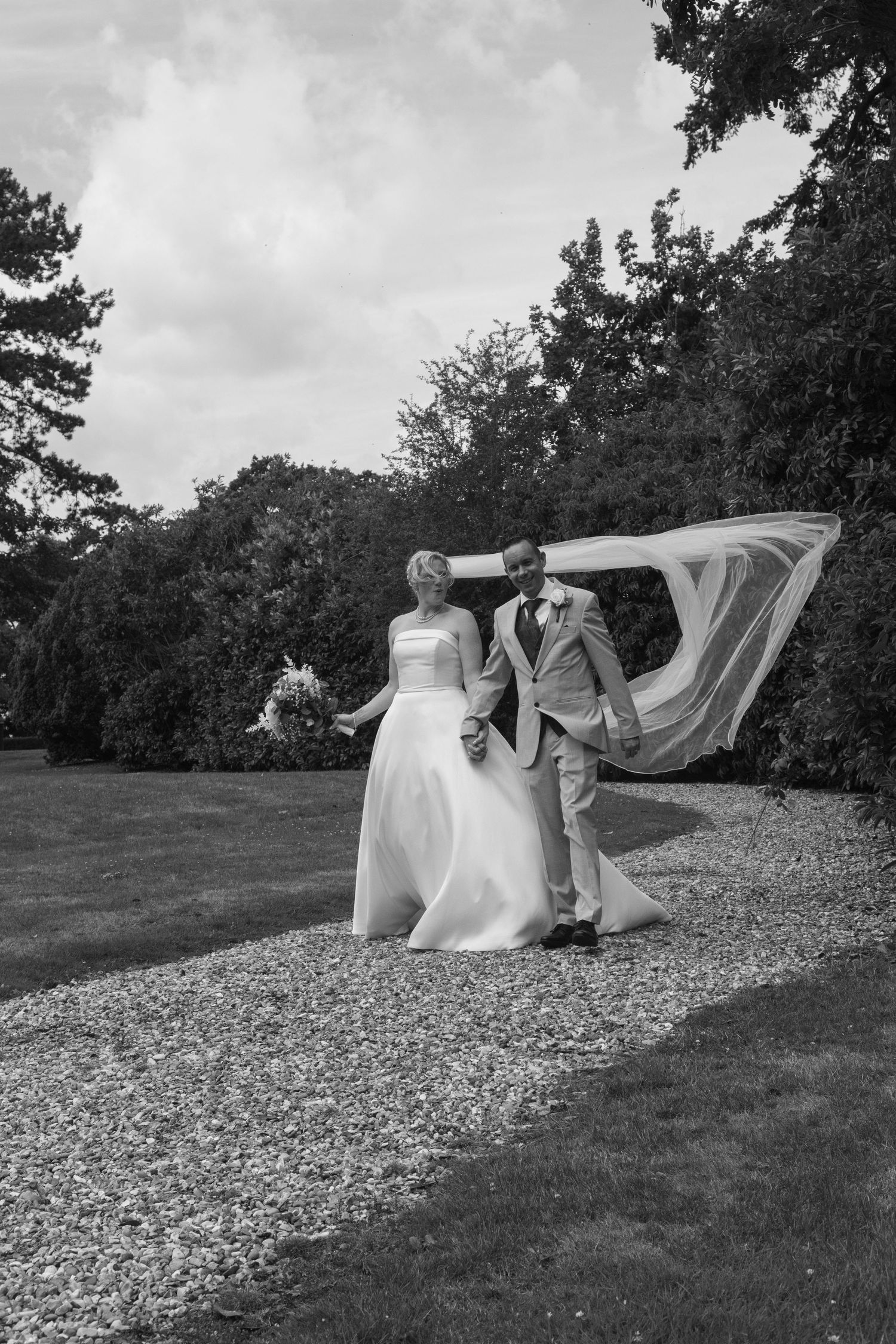 Black and white photo of bride and groom walking on gravel path with veil blowing in the wind.