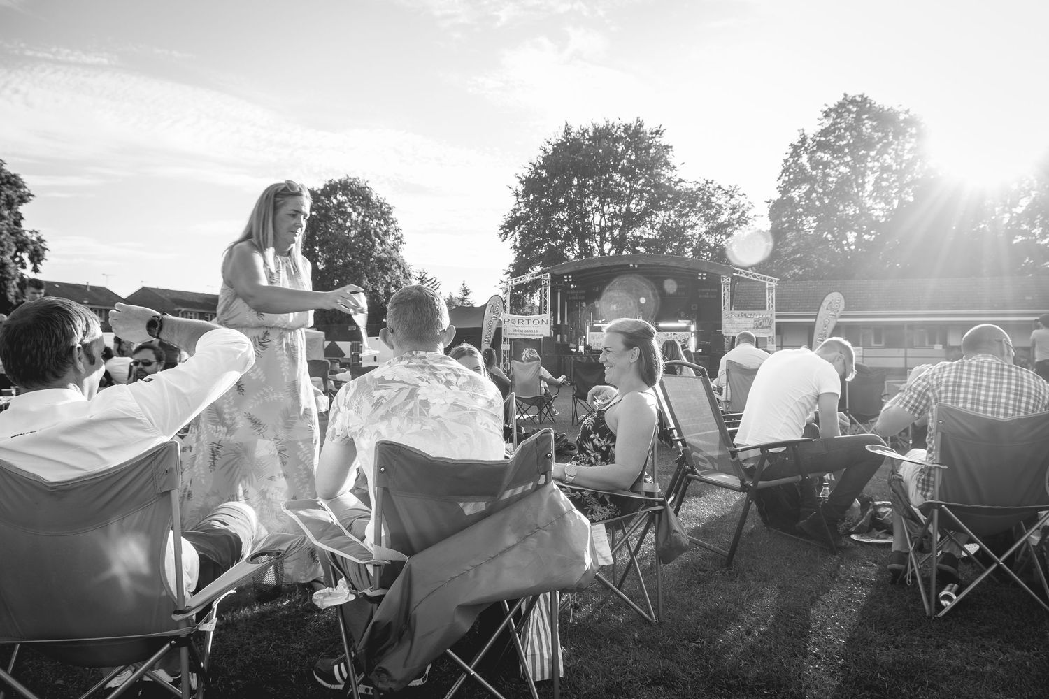Black and white photo of people relaxing in folding chairs at an outdoor summer event with sunlight filtering through trees.
