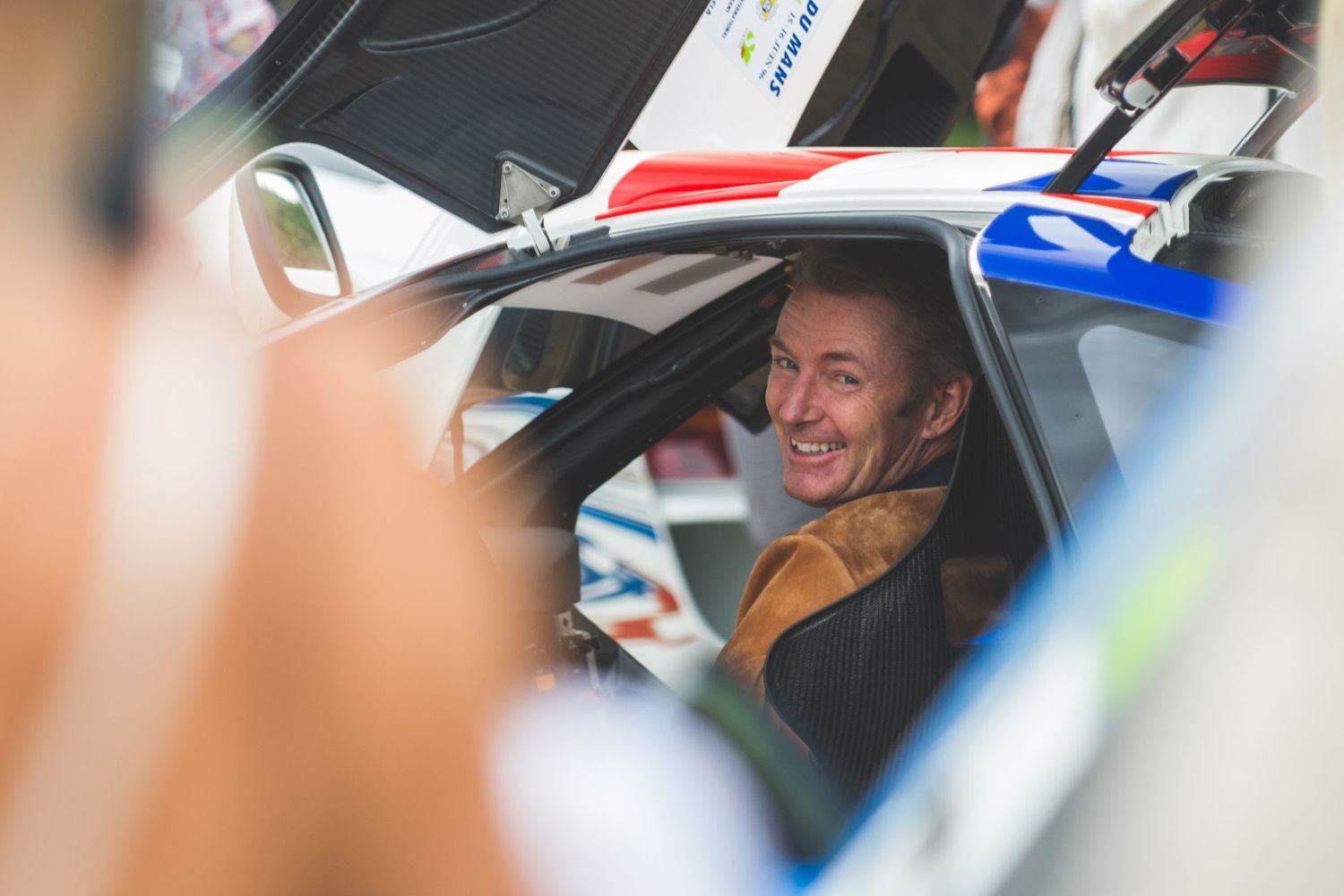A smiling Simon Kidston sits in the cockpit of a modern race car at a motorsport event.