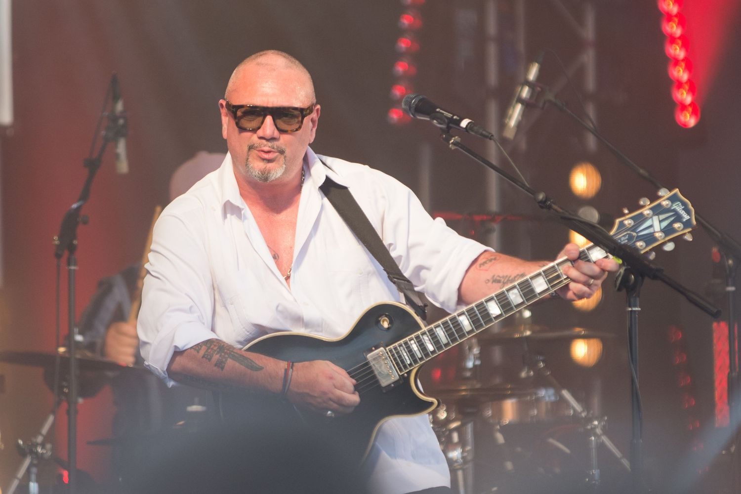 Fun Lovin' Criminals front man Heuey Morgan in a white shirt plays black Les Paul guitar on outdoor stage.