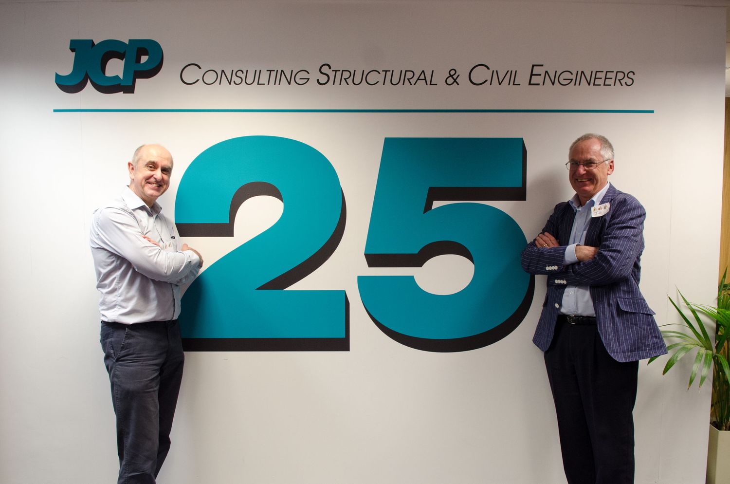 Two men posing beside a large 25th anniversary sign for JCP Consulting Structural and Civil Engineers.