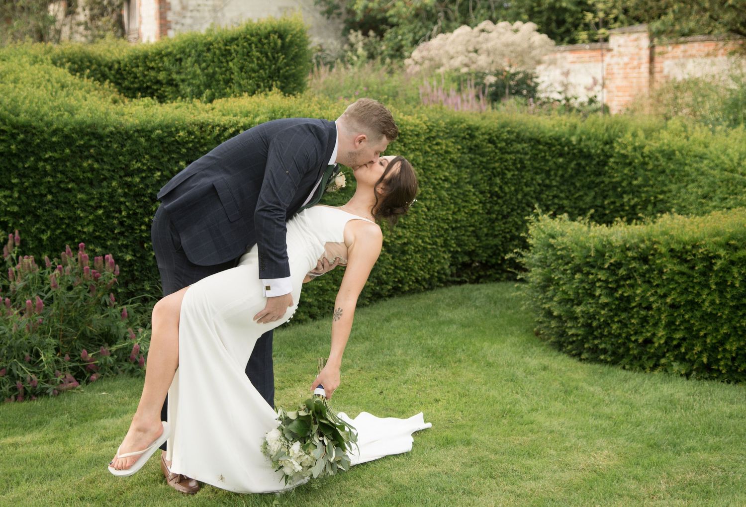 Groom in navy suit dips bride in sleek white gown for a romantic kiss on a manicured garden lawn.