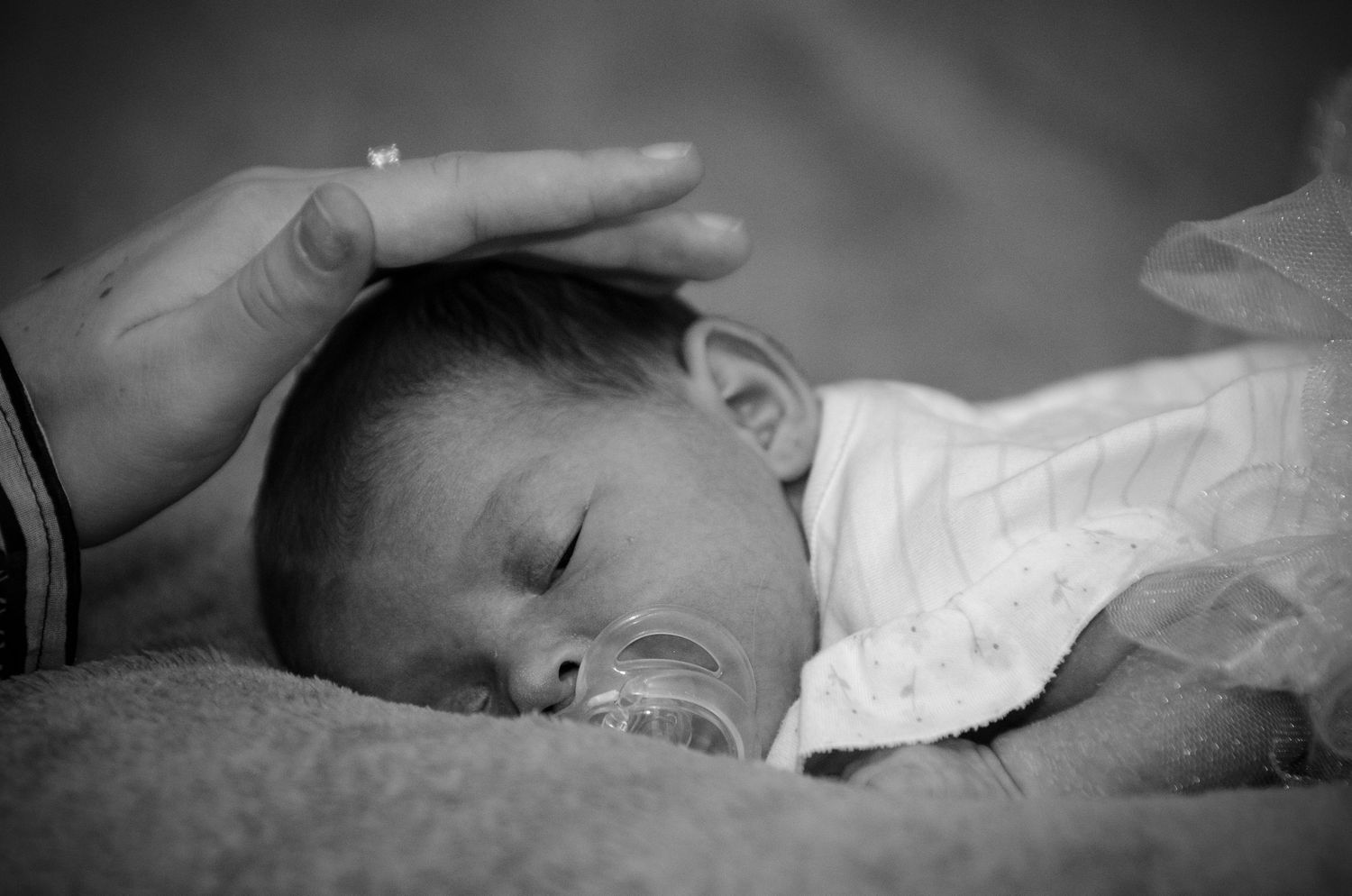Black and white photo of a sleeping newborn baby with a pacifier, gently cradled by an adult hand.