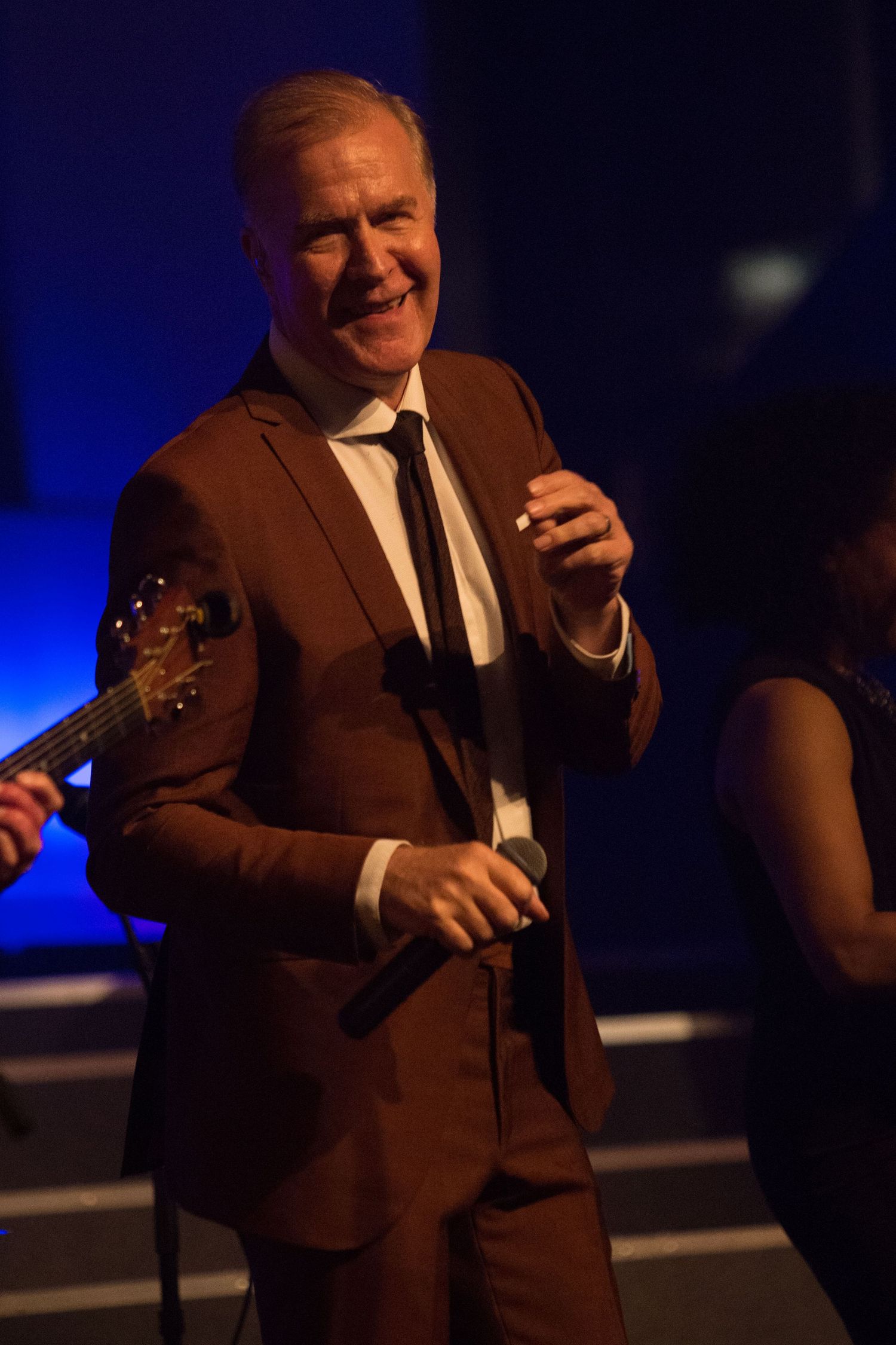 ABC's Martin Fry dressed in a brown suit smiling and holding microphone on stage with guitarist performing behind him.