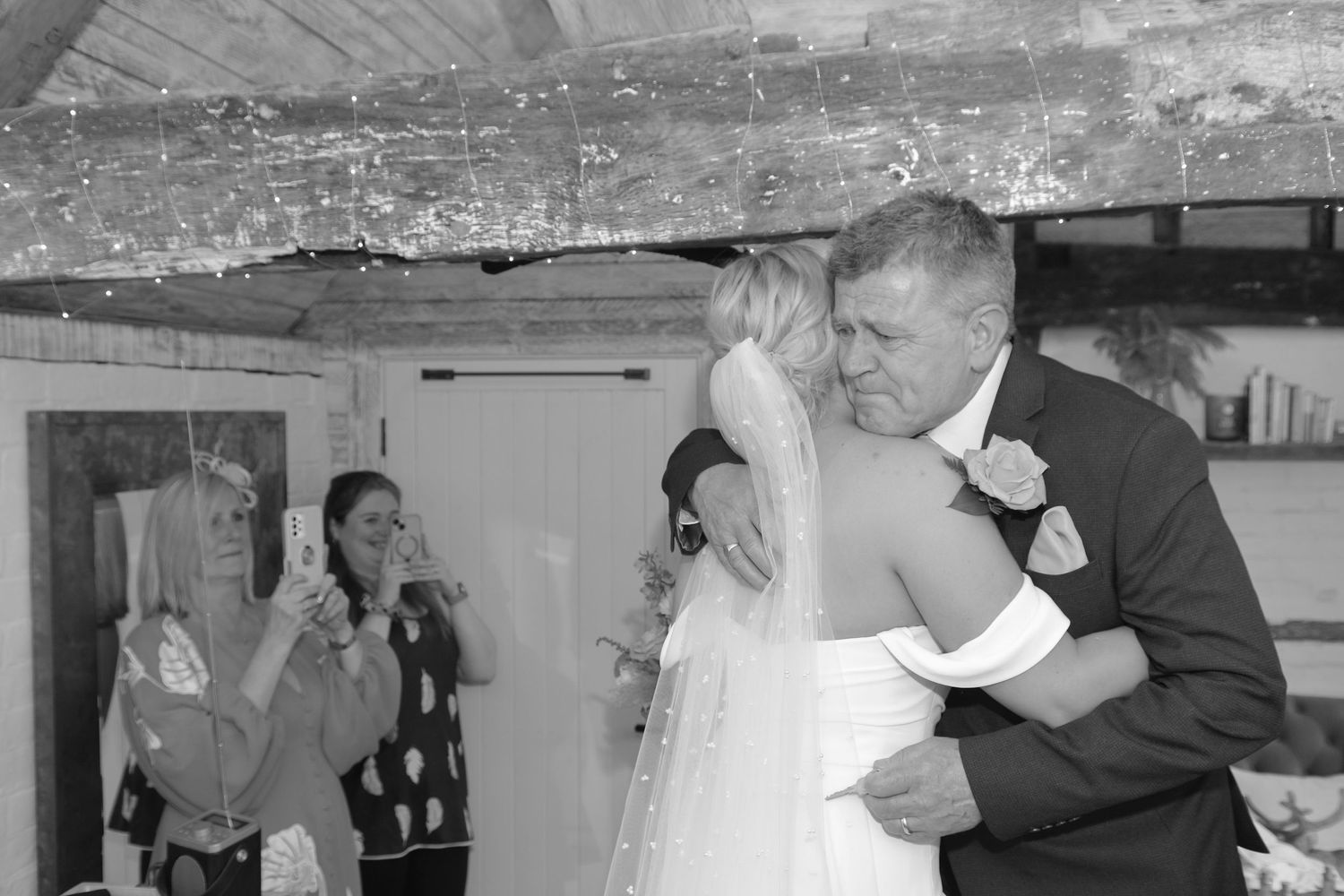 Emotional black and white wedding photo of bride embracing father with guests watching nearby.