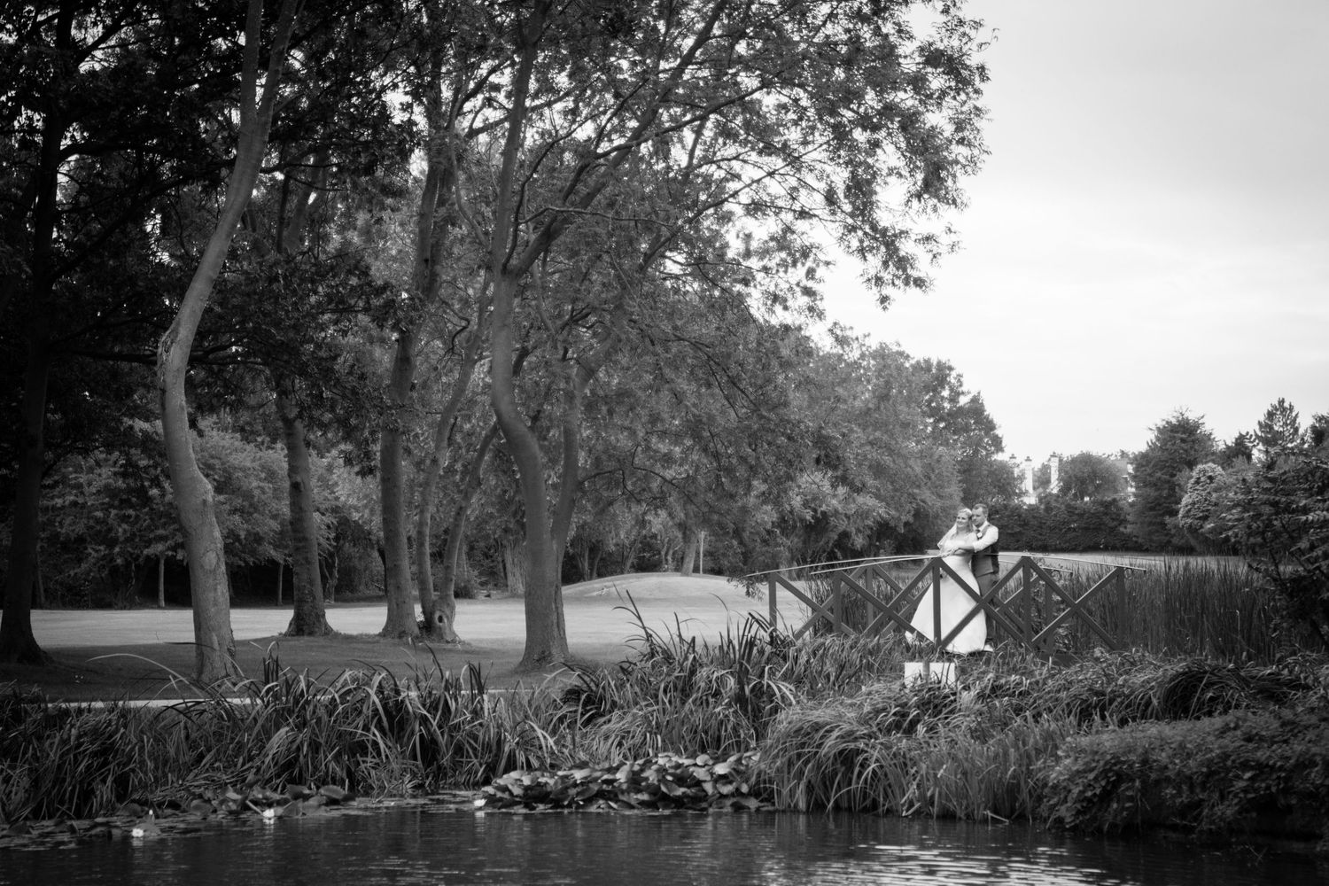 Black and white romantic photo of a couple on a bridge surrounded by trees and water.