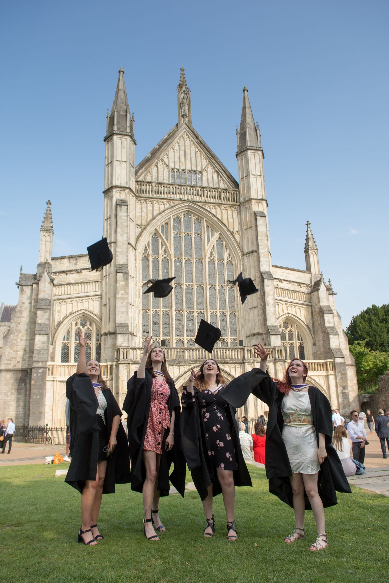 Four female graduates in black gowns toss mortarboard caps in front of a Gothic cathedral on a sunny day.