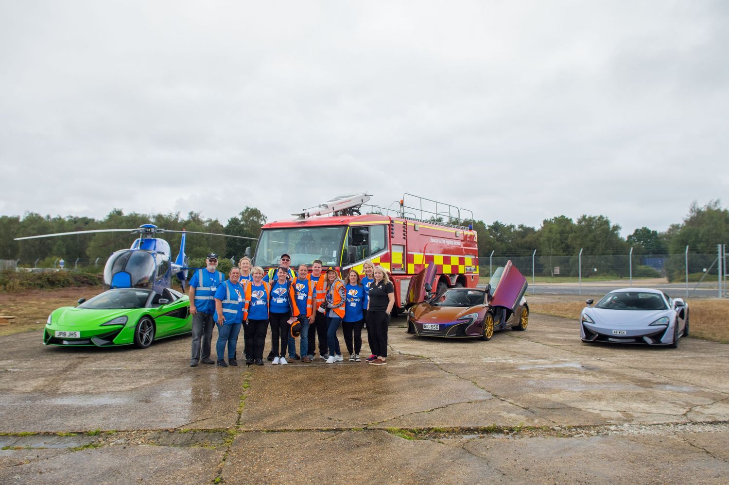 Group of people posing with McLaren supercars, fire truck, and helicopter on an airfield on a cloudy day.