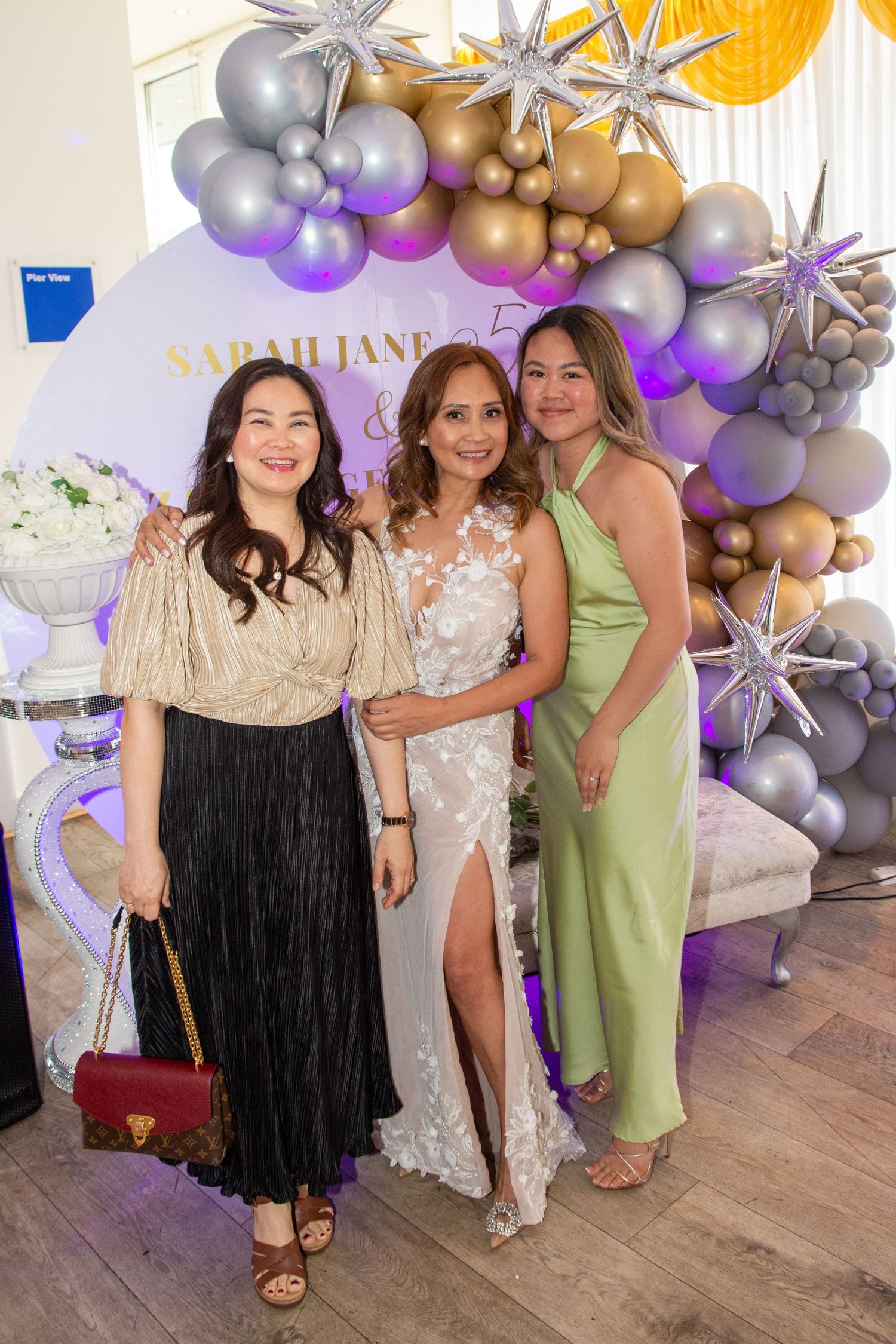 Three women posing together at a birthday celebration with a decorative balloon arch backdrop.