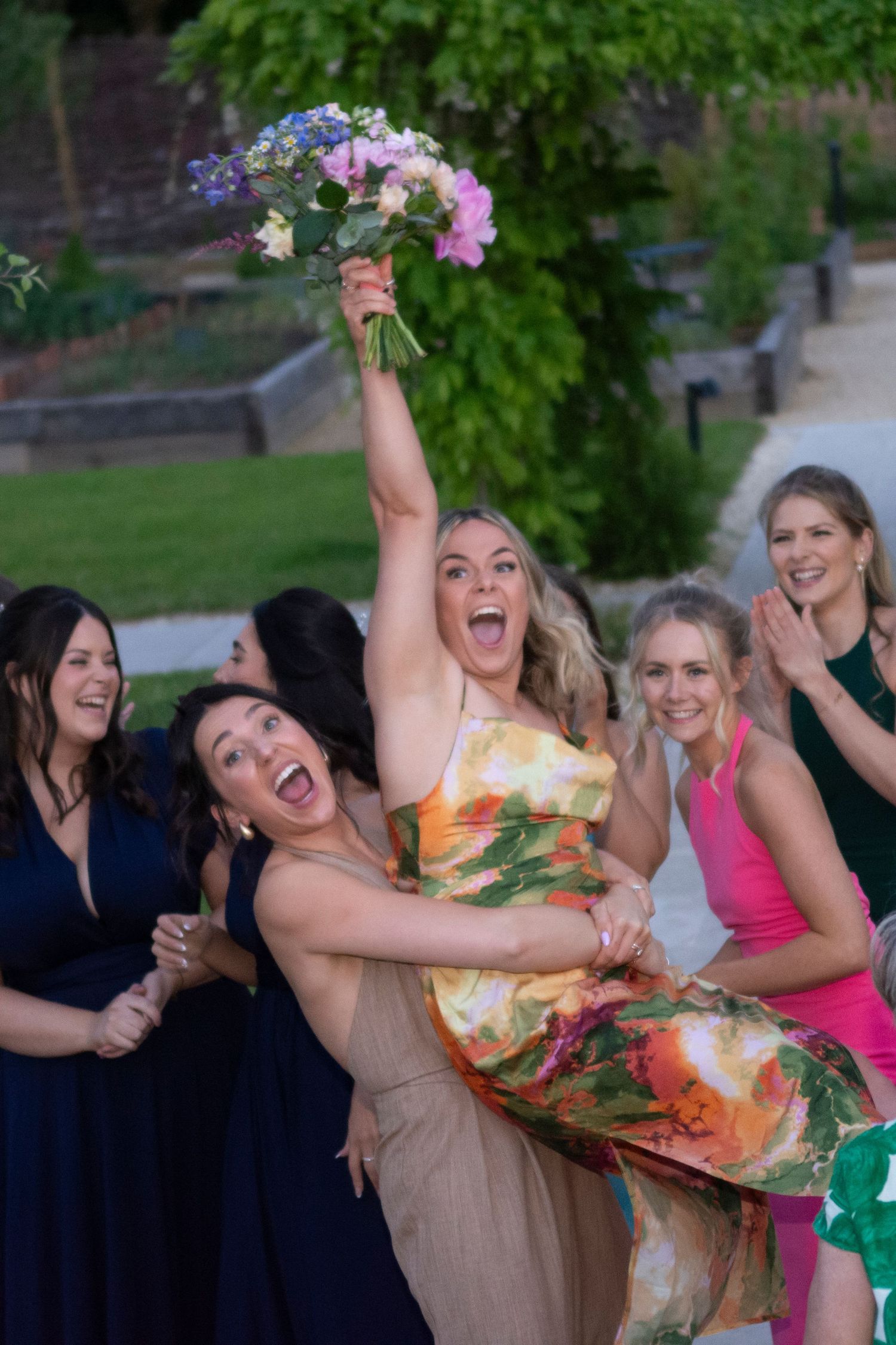 Excited woman catches bridal bouquet surrounded by cheering guests in colorful dresses at outdoor wedding celebration.