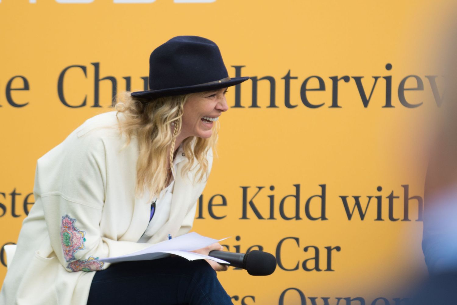 Jodie Kidd in a black hat speaks at an interview panel event with a yellow branded backdrop.