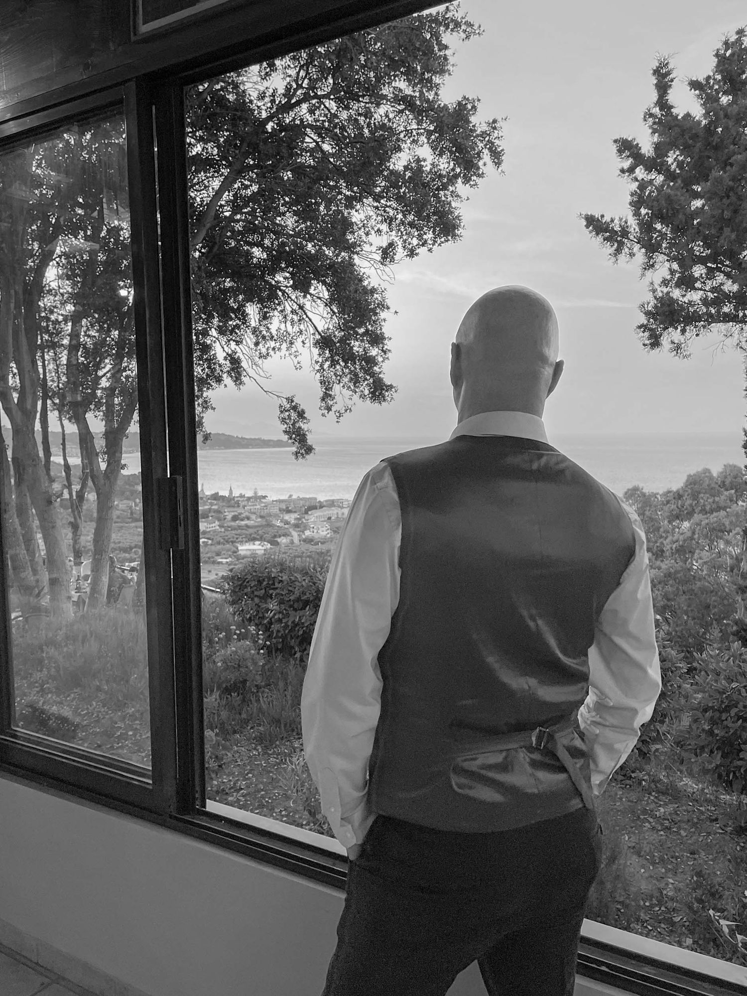 Bald groom in vest and white shirt gazing out large window at scenic coastal landscape in black and white photo.