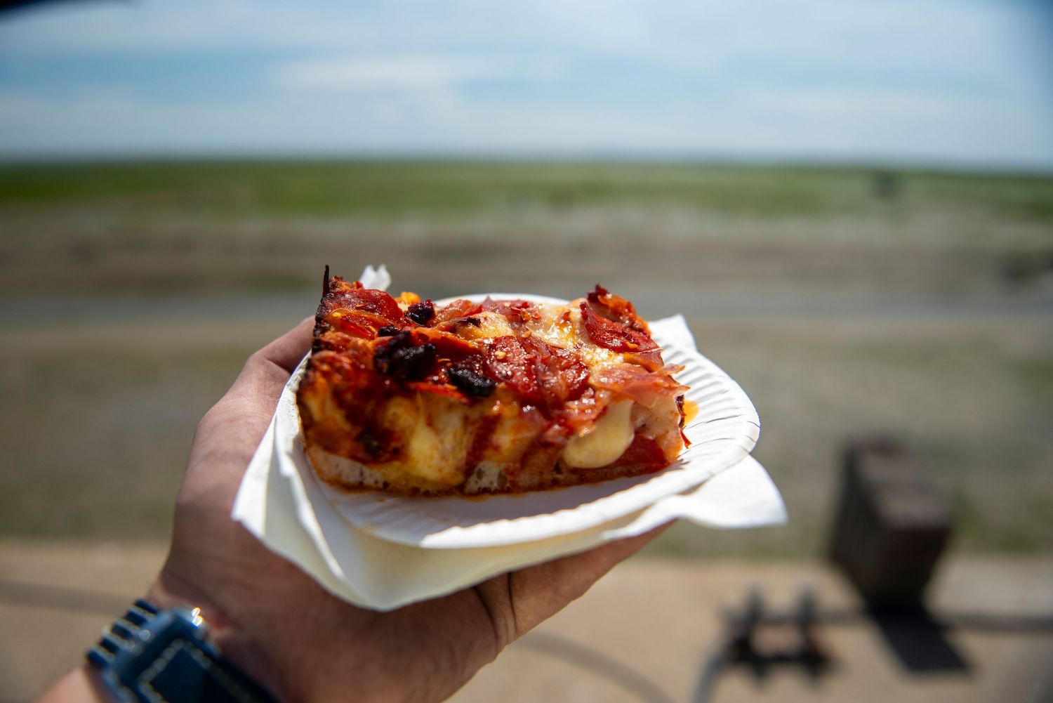 A hand holds a paper plate with a slice of pizza topped with tomato sauce and caramelized onions outdoors.