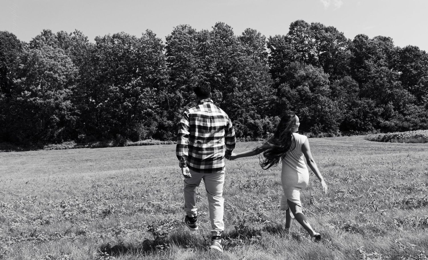 Black and white photo of a couple holding hands walking through a grassy meadow with trees.