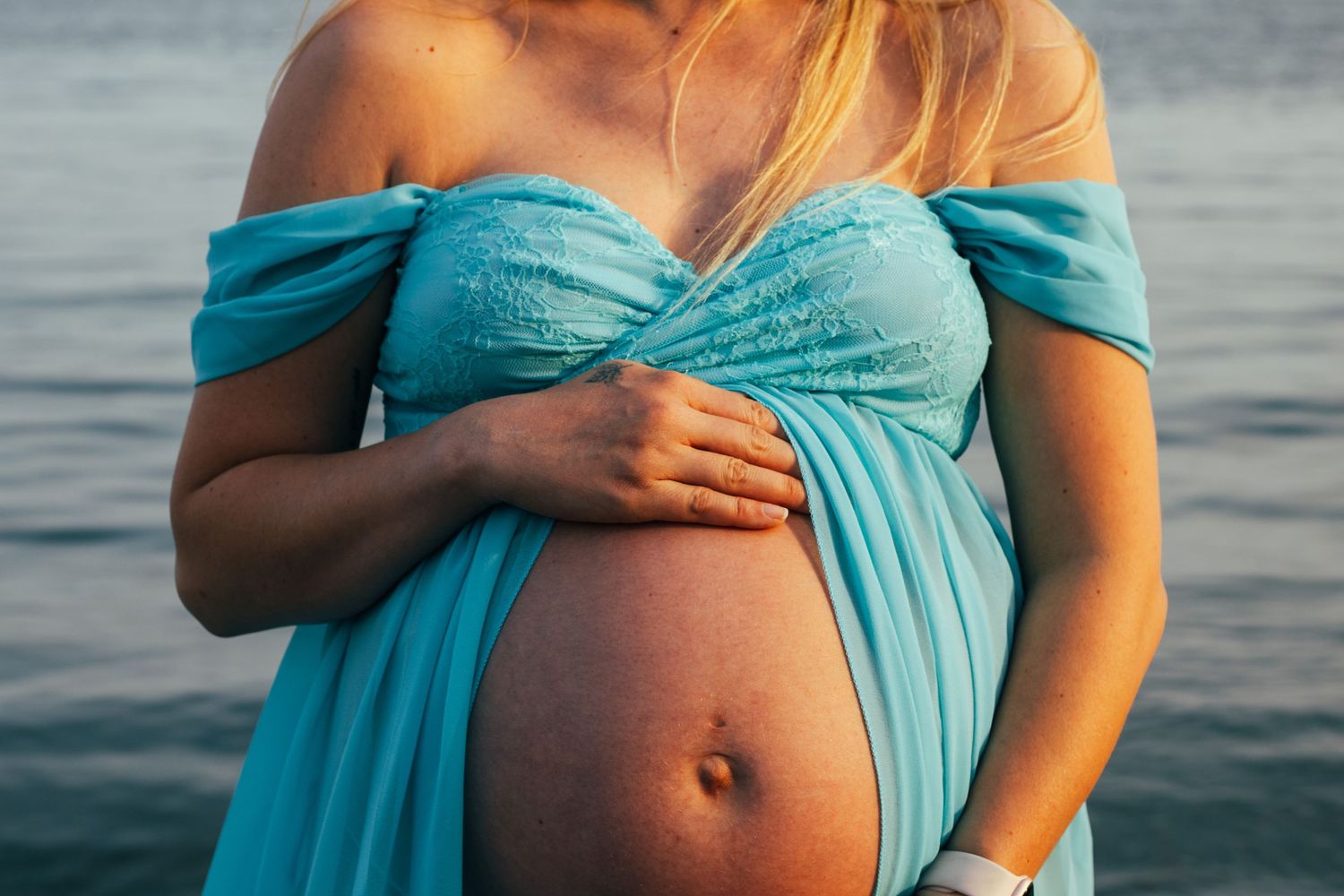 Pregnant woman in teal off-shoulder gown cradling her baby bump near the ocean at sunset.