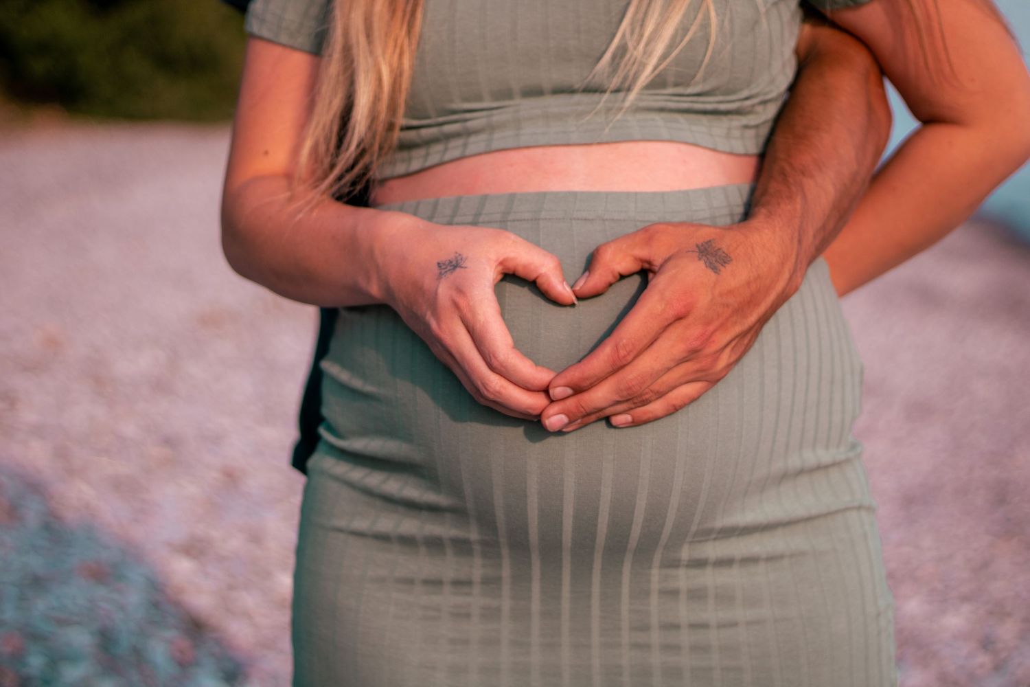 Pregnant woman in sage green outfit forming heart shape with hands over baby bump outdoors.