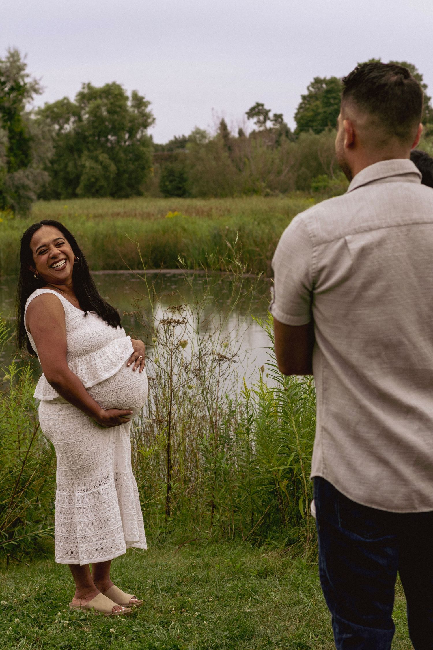 Smiling pregnant woman in white dress laughing with partner near a serene pond in a lush green park.