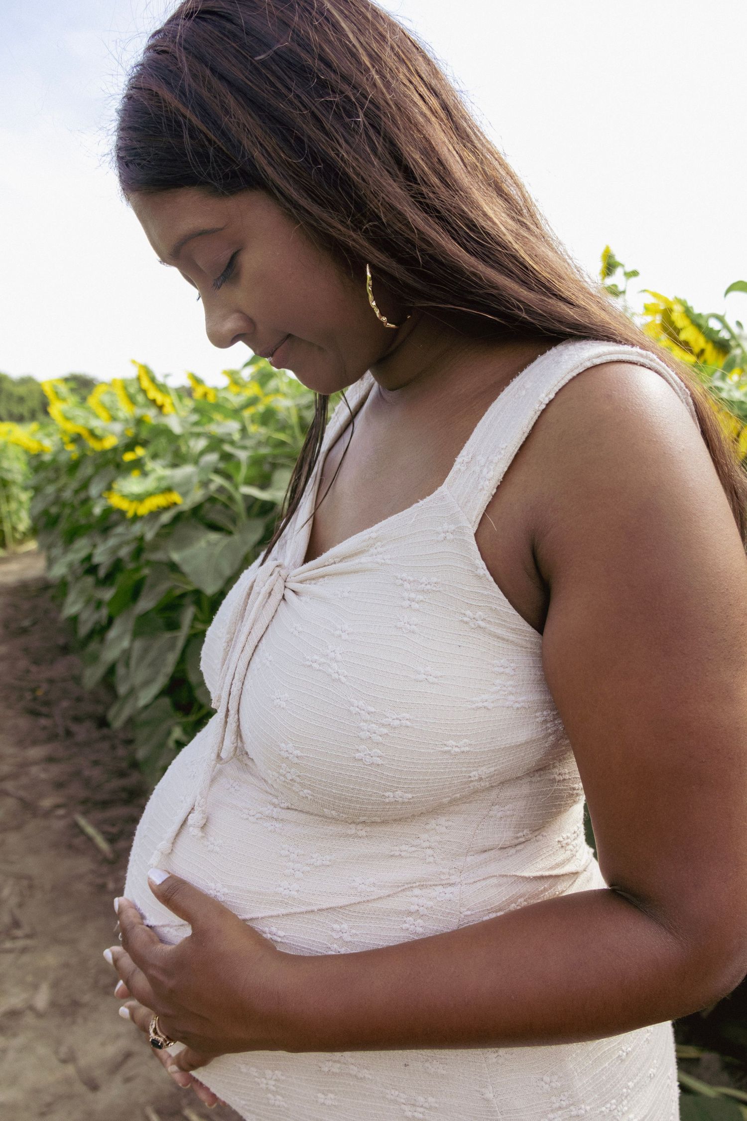 Pregnant woman in white dress cradling her baby bump in a sunflower field.