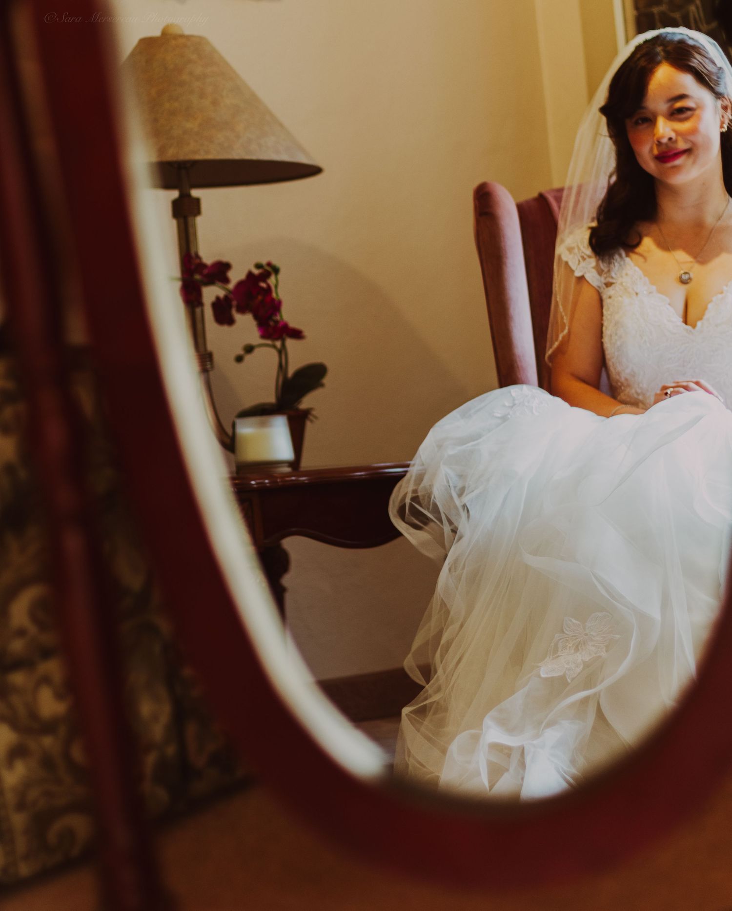 Bride in white wedding gown and veil sitting in a chair, reflected in an oval mirror with a lamp and orchid nearby.